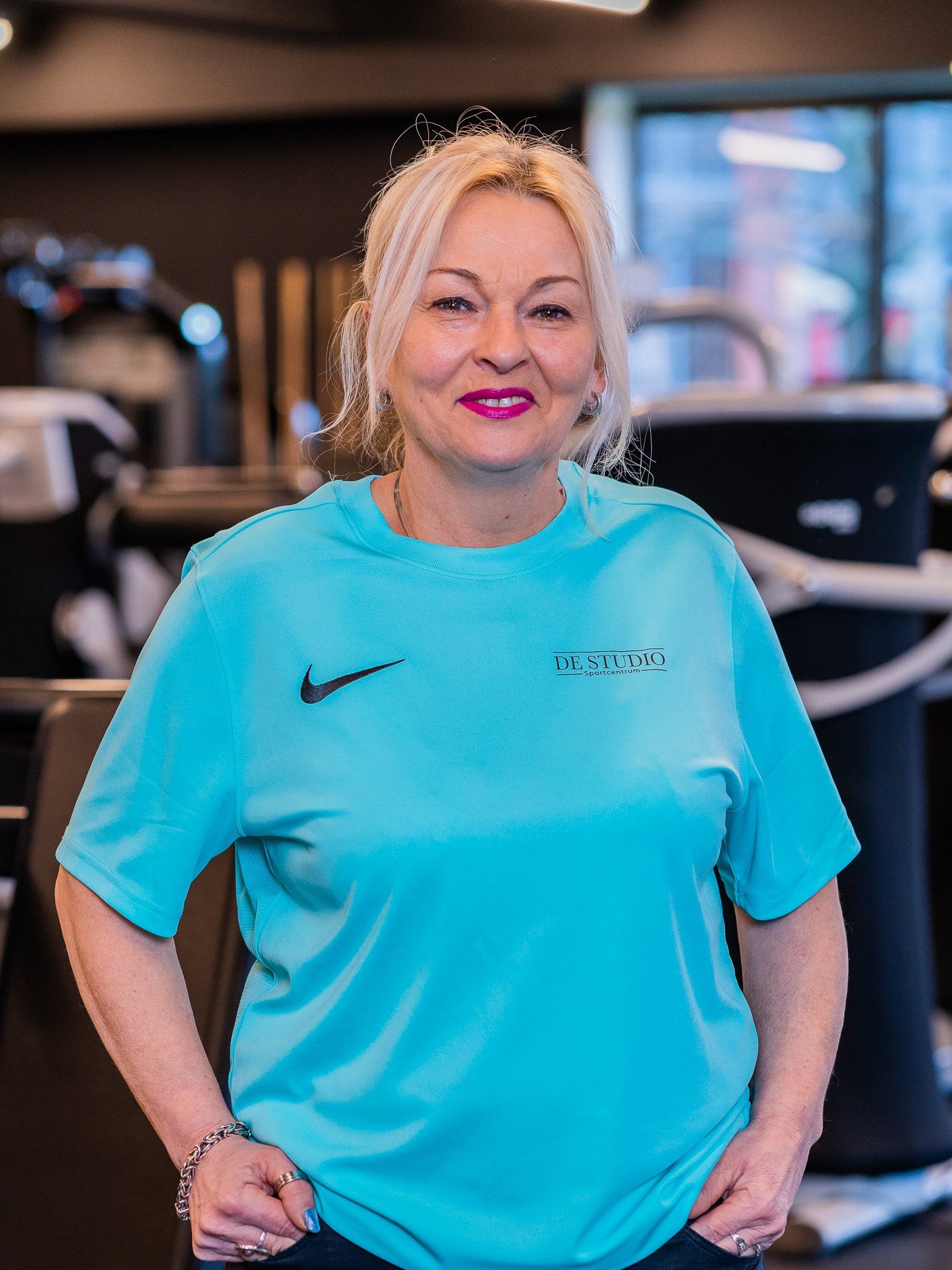 Een vrouw in een blauw Nike-shirt staat in een sportschool met haar handen op haar heupen.