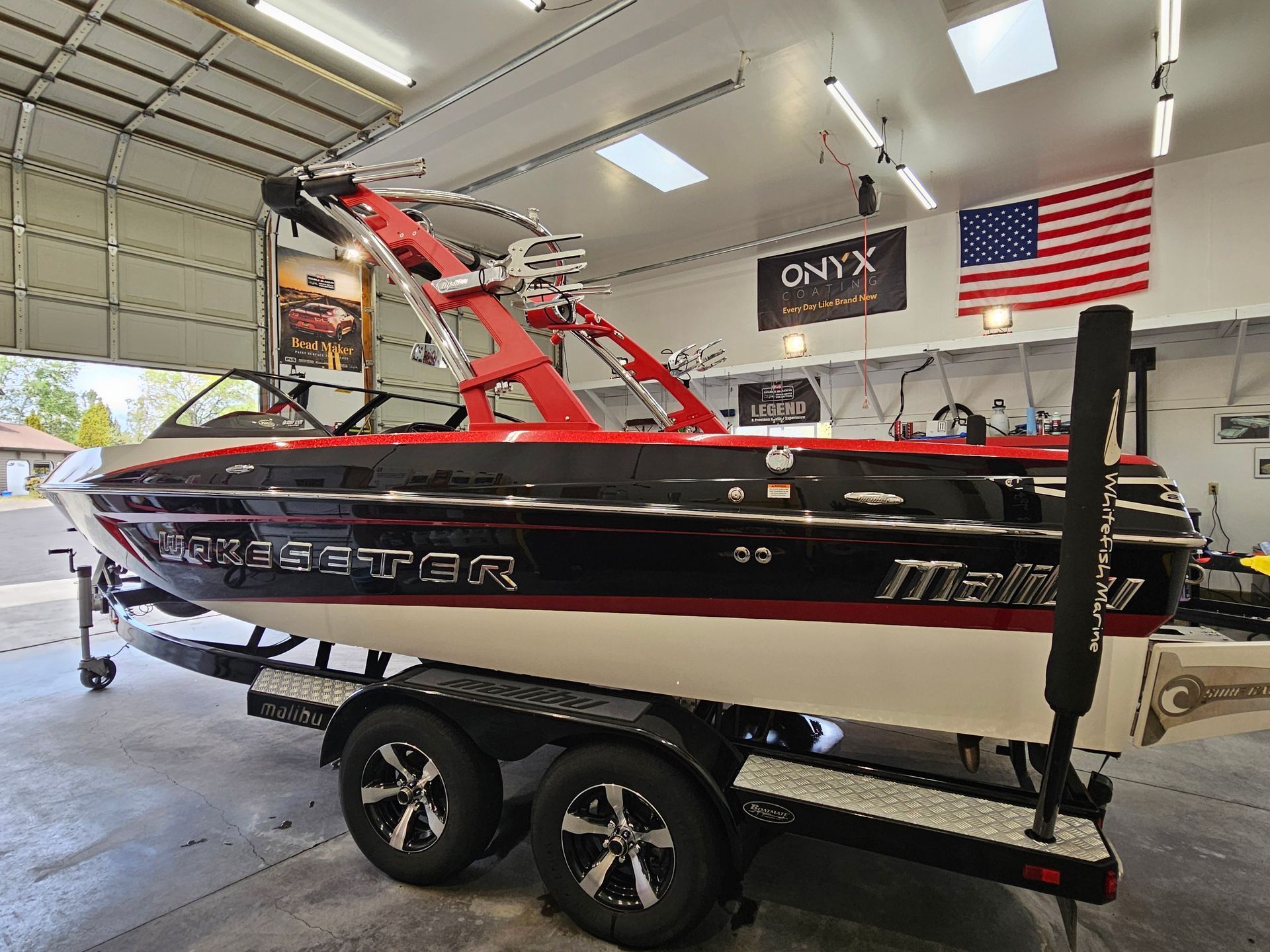 Black and red Malibu boat on a trailer, with a red wakeboard tower, parked indoors with an American flag.