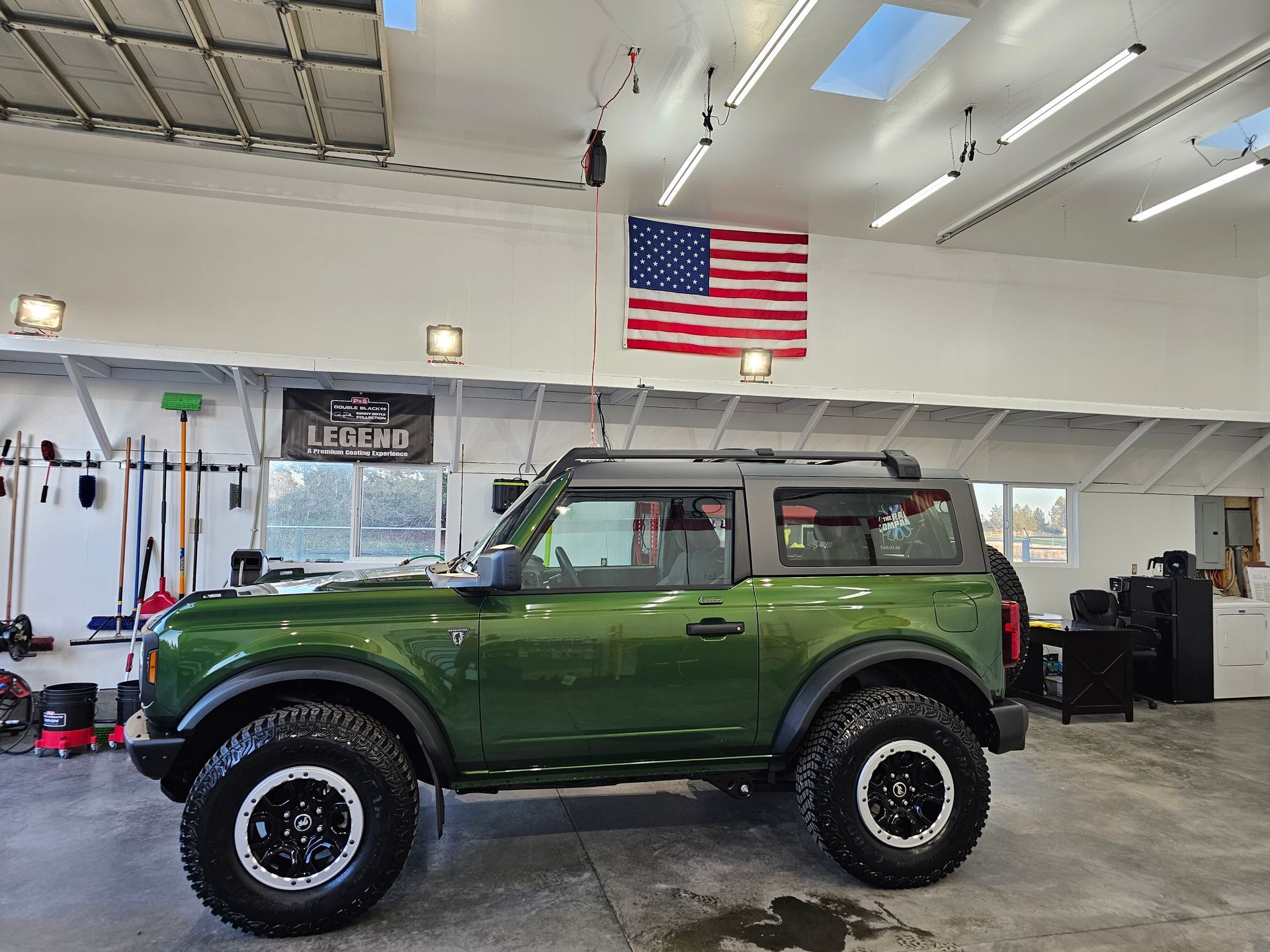 Green Ford Bronco with black wheels parked inside a garage, American flag displayed on the wall.