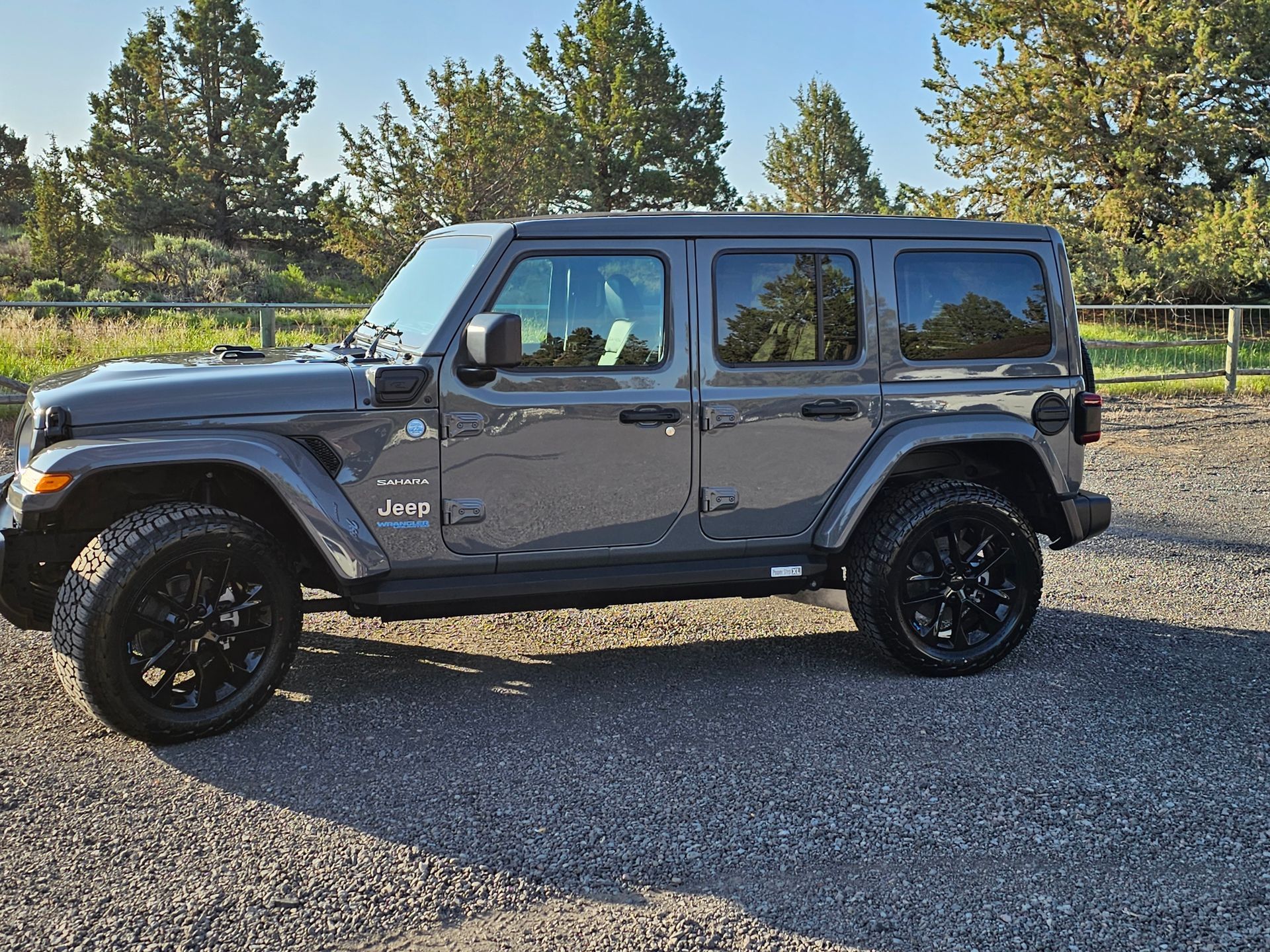 Gray Jeep Wrangler parked on gravel. Black wheels, tinted windows, sunny day.