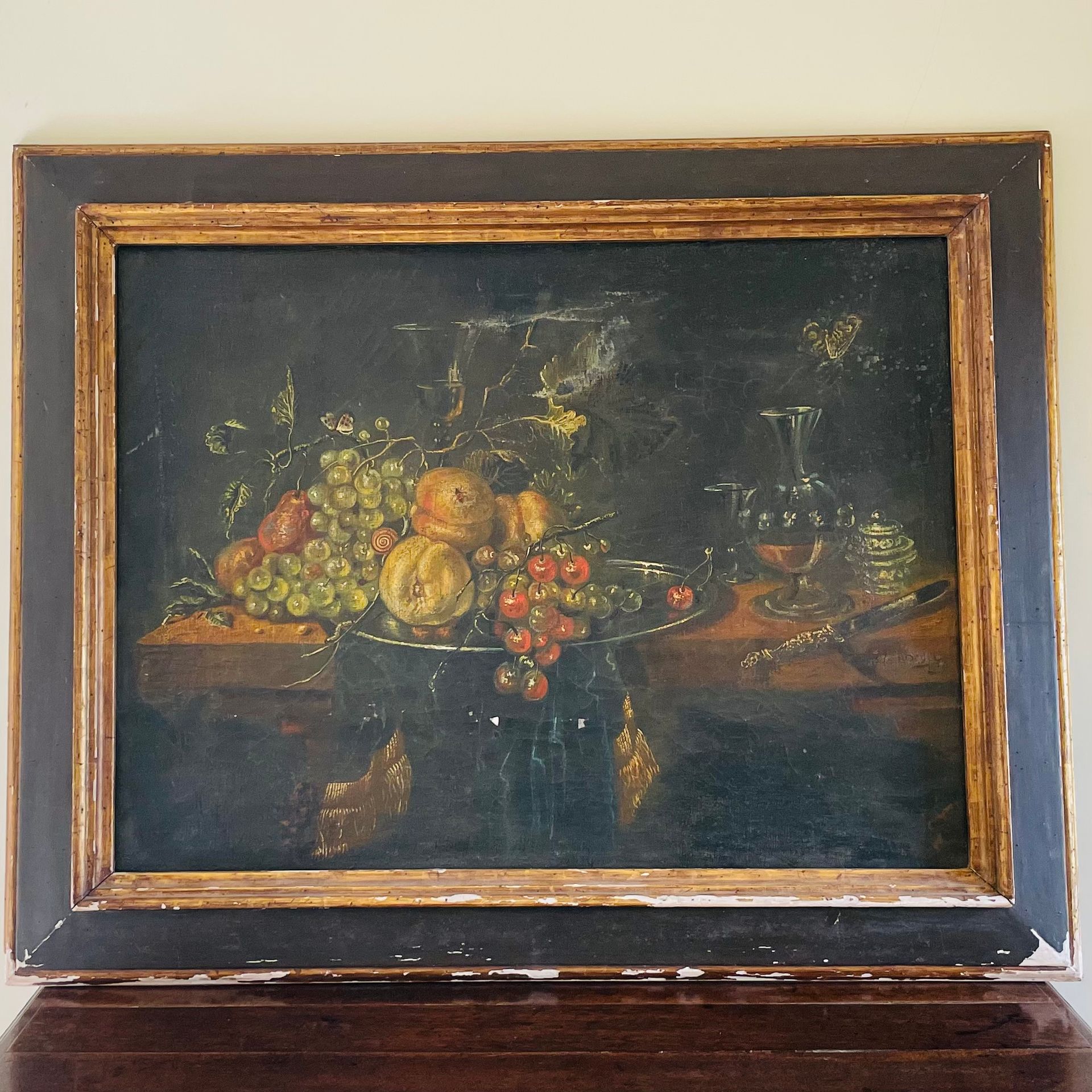 A still life painting which depicts decadent fruit on an elaborate platter, a knife tumbling from a dressed table, and several insects including a butterfly.