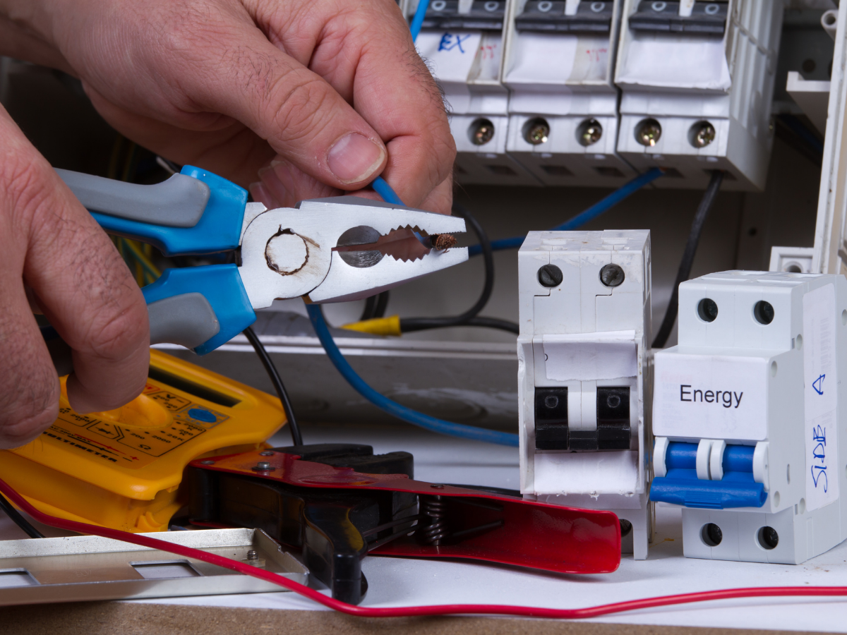 Electrician using pliers on a wire near circuit breakers, indoors.