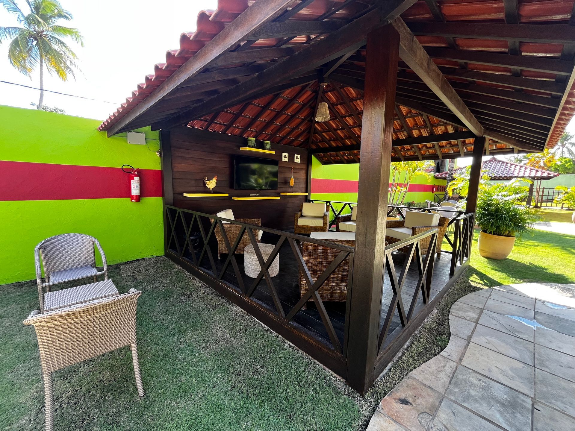Gazebo de madeira com telhado de telha, área de estar e televisão contra uma parede verde vibrante com uma faixa vermelha.
