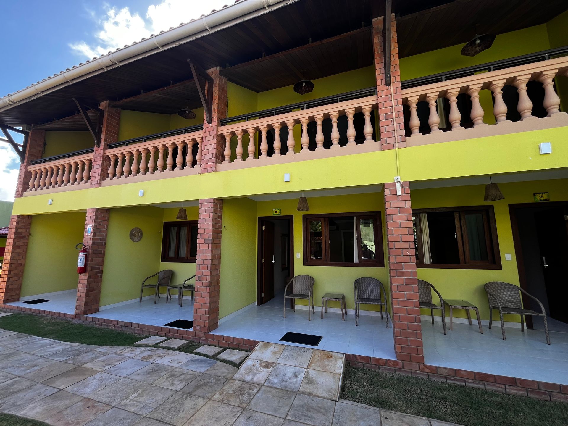 Prédio amarelo de dois andares com colunas de tijolos e varandas, área externa com mesas e cadeiras, gramado verde.