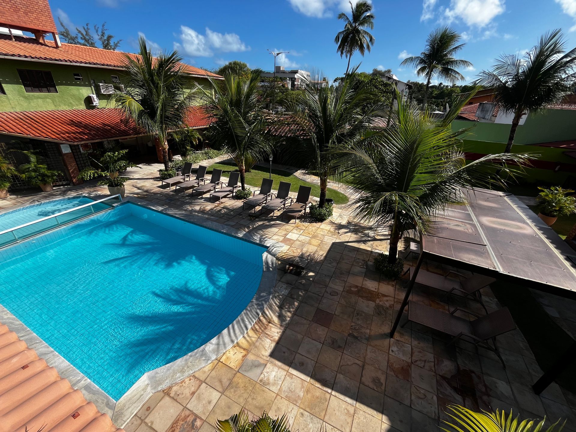 Área da piscina com água azul, palmeiras, espreguiçadeiras e edifícios com telhados de telha vermelha.