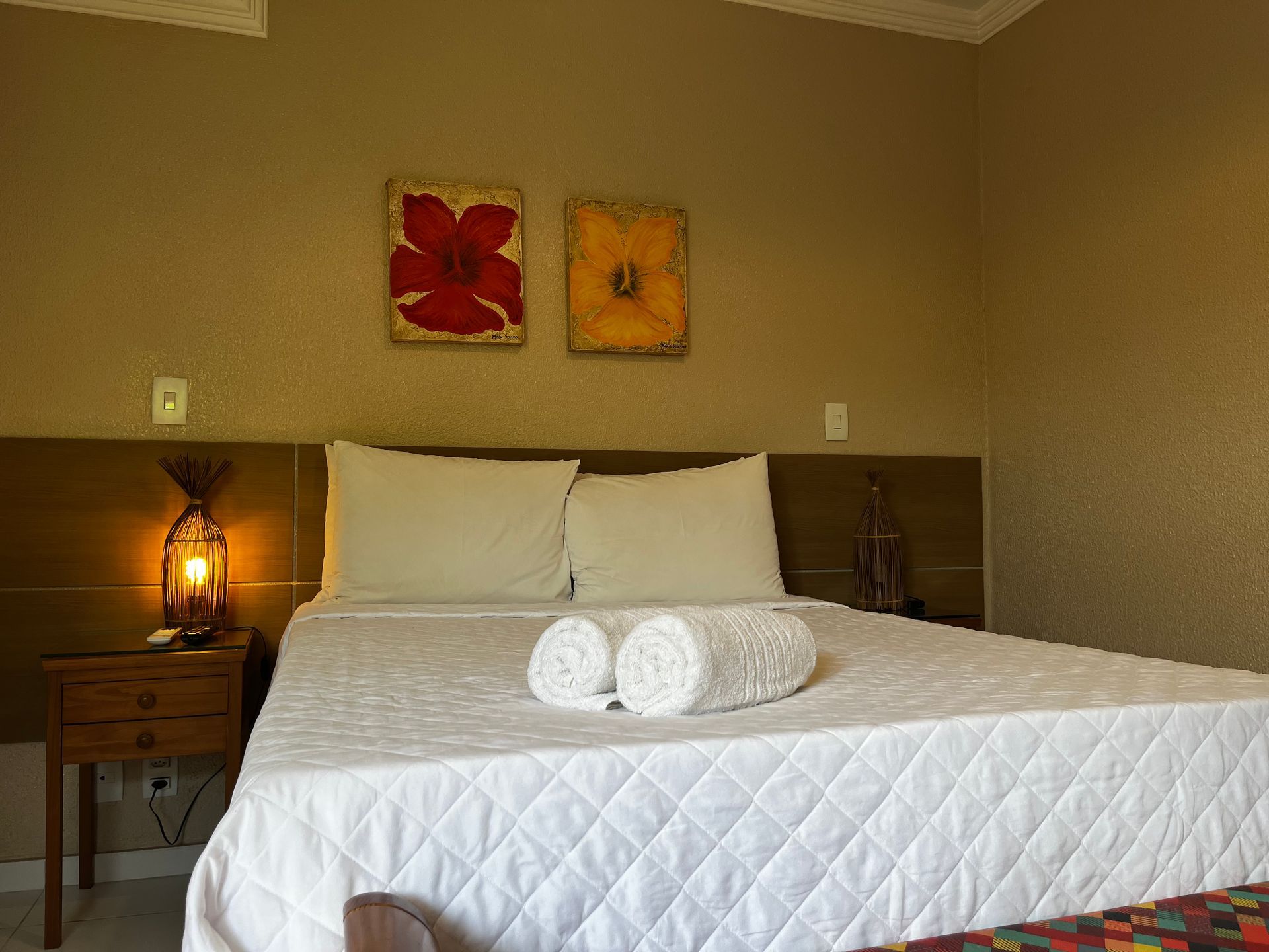 Quarto com cama, mesa de cabeceira e obras de arte na parede. A cama tem roupa de cama branca e duas toalhas enroladas.
