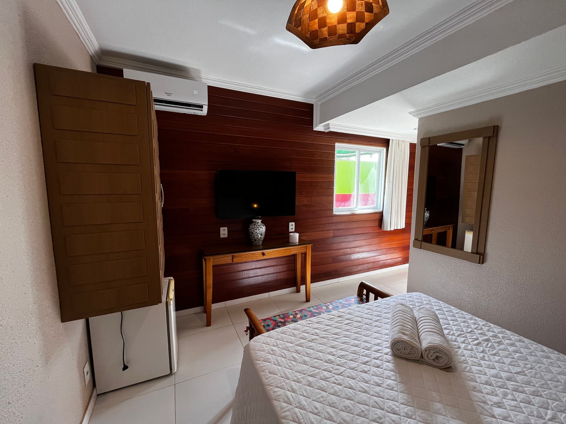 Quarto com painéis de madeira, TV, cama com toalhas e janela.