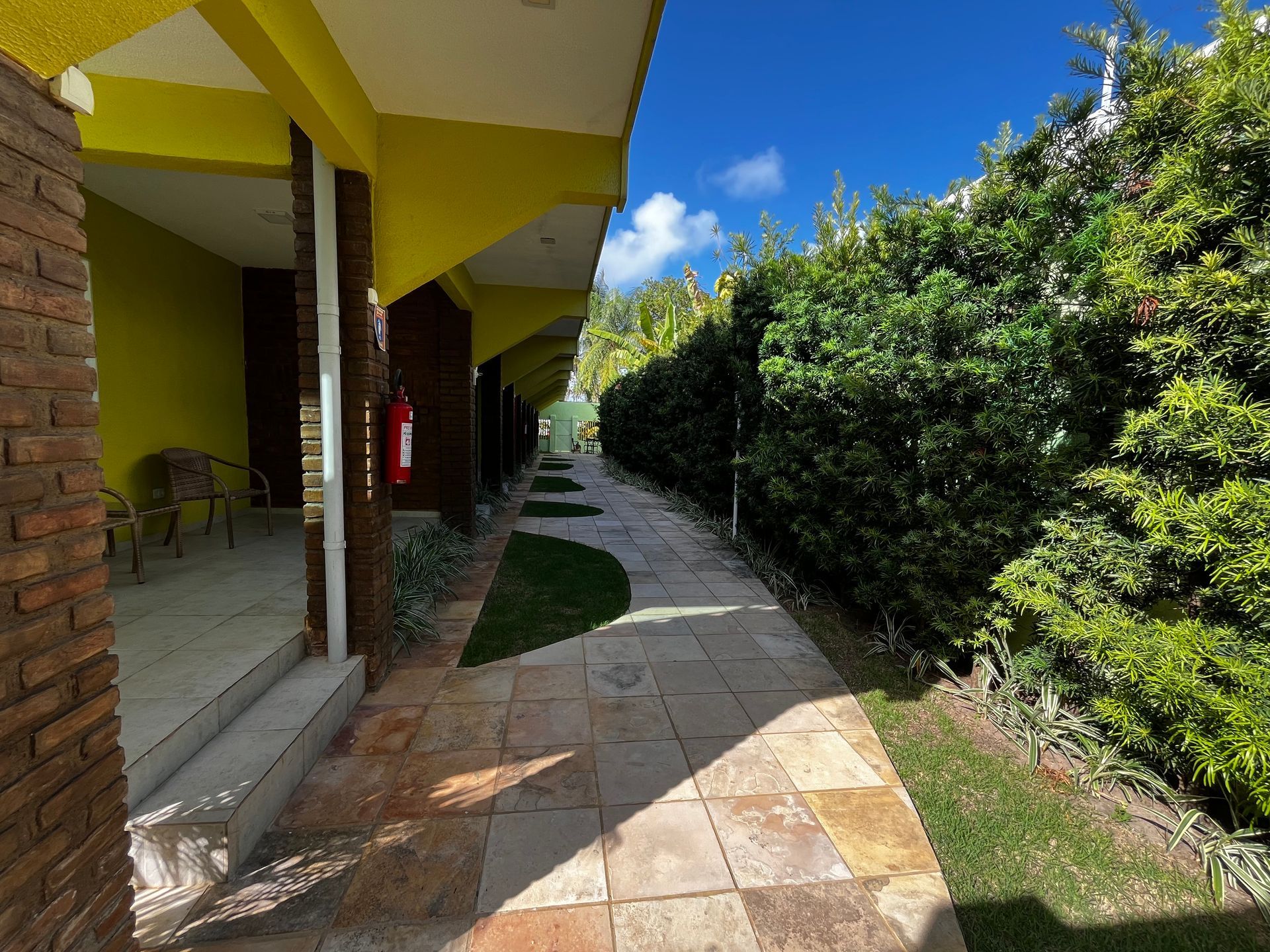 Passarela entre um prédio com detalhes em amarelo e uma cerca viva em um dia ensolarado.