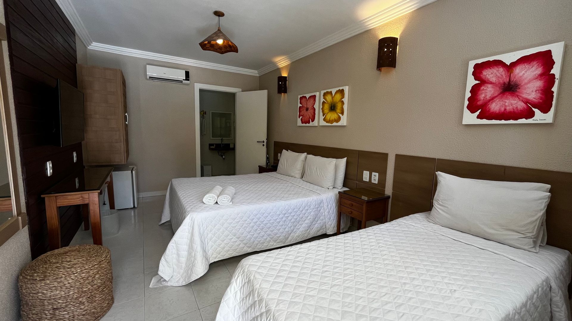 Quarto de hotel com duas camas, obras de arte e um pequeno frigobar. Cores neutras e iluminação de teto.