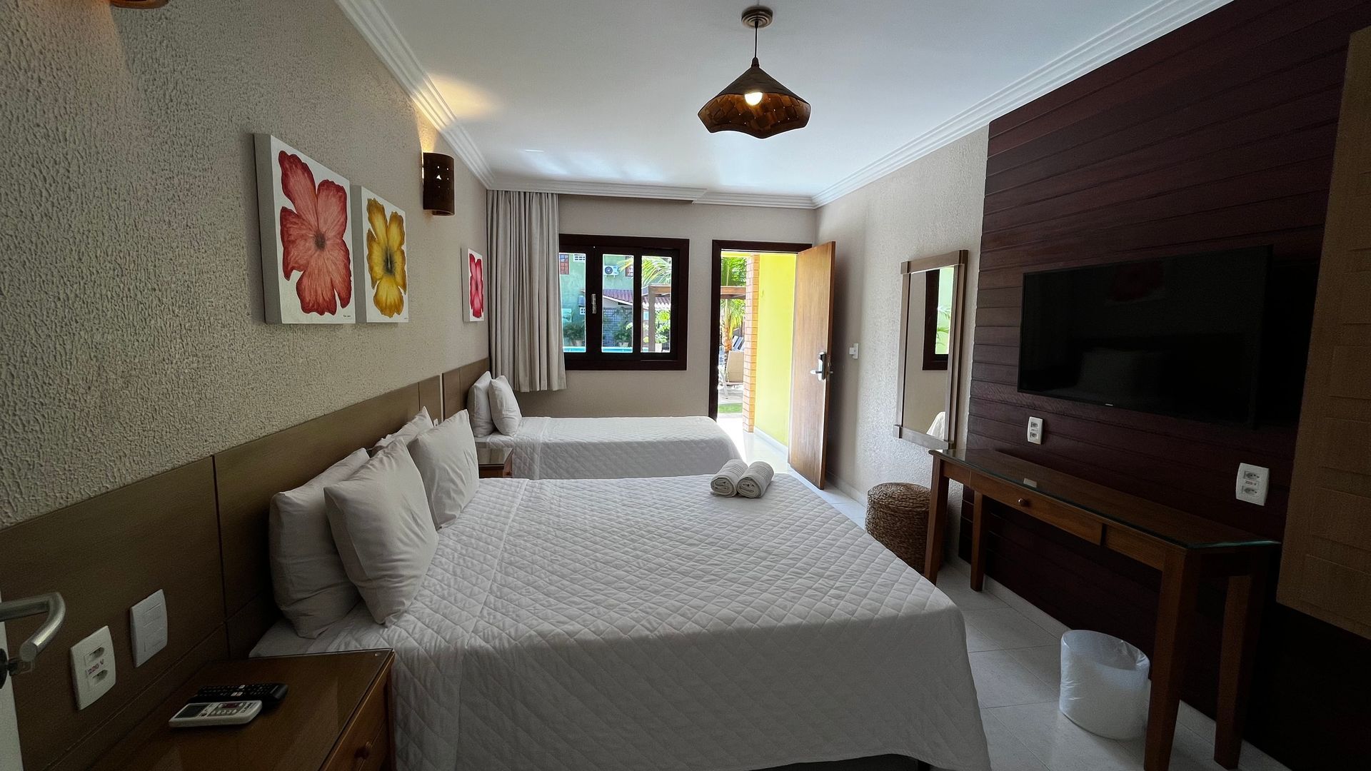 Quarto de hotel com duas camas, TV, escrivaninha e porta aberta para o exterior. Cores claras e neutras.