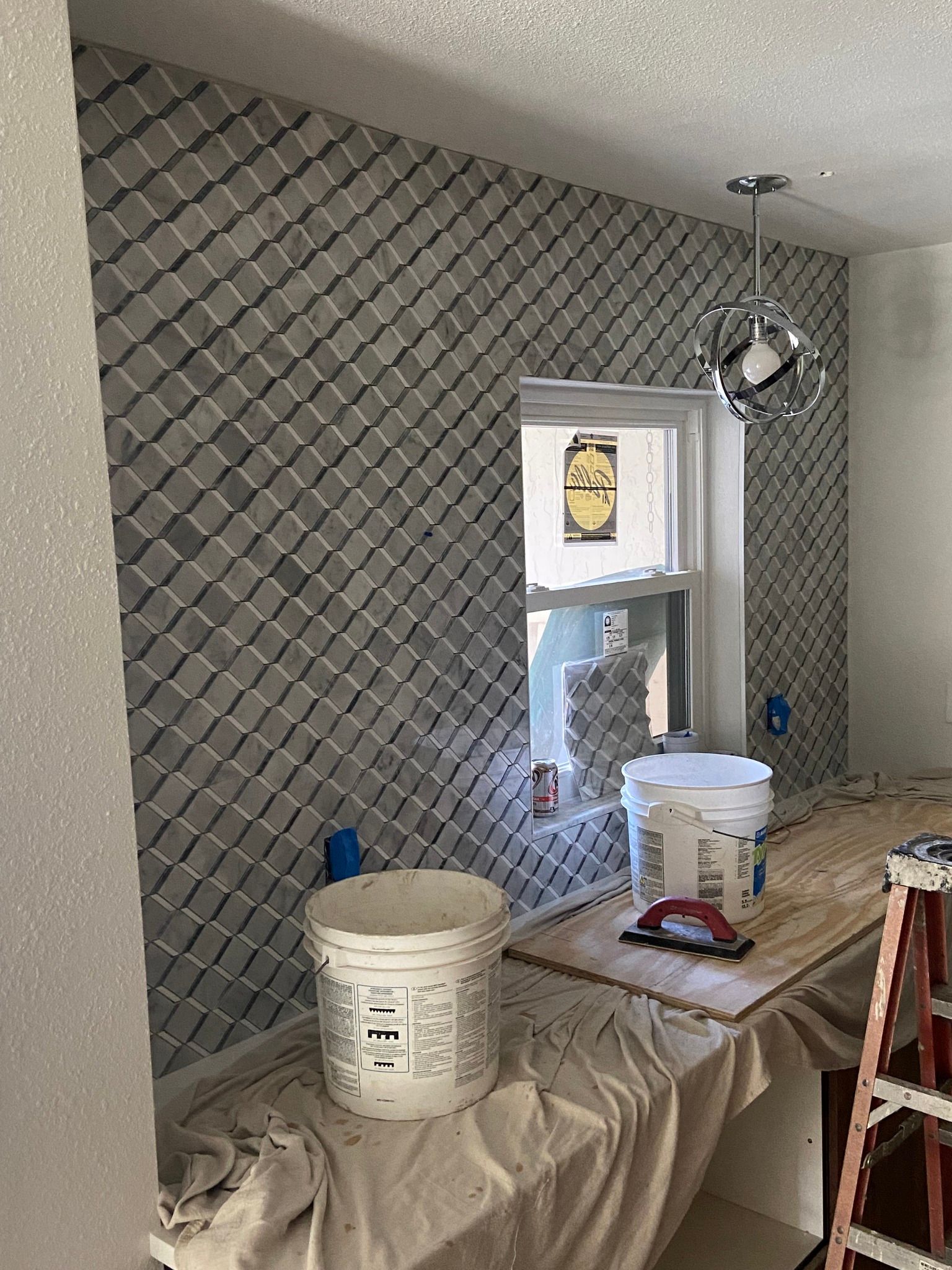 Tiled wall in progress with gray diamond tiles, window, buckets, and a partially constructed counter.