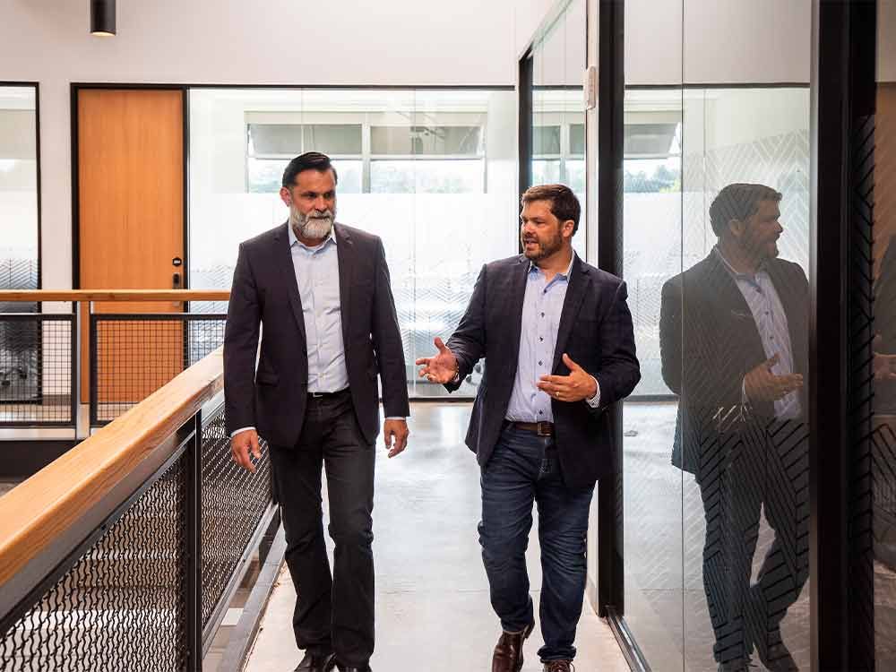Two men walking in a modern office hallway, talking beside glass walls and a railing.