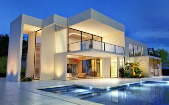 Modern white house with glass walls, balcony, and pool, illuminated at dusk.