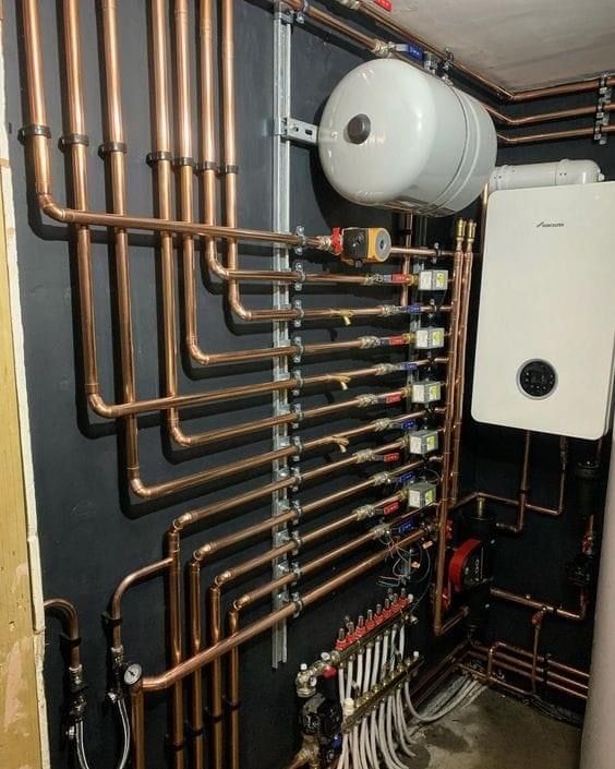 Copper pipes and a boiler system installed on a black wall in a mechanical room.