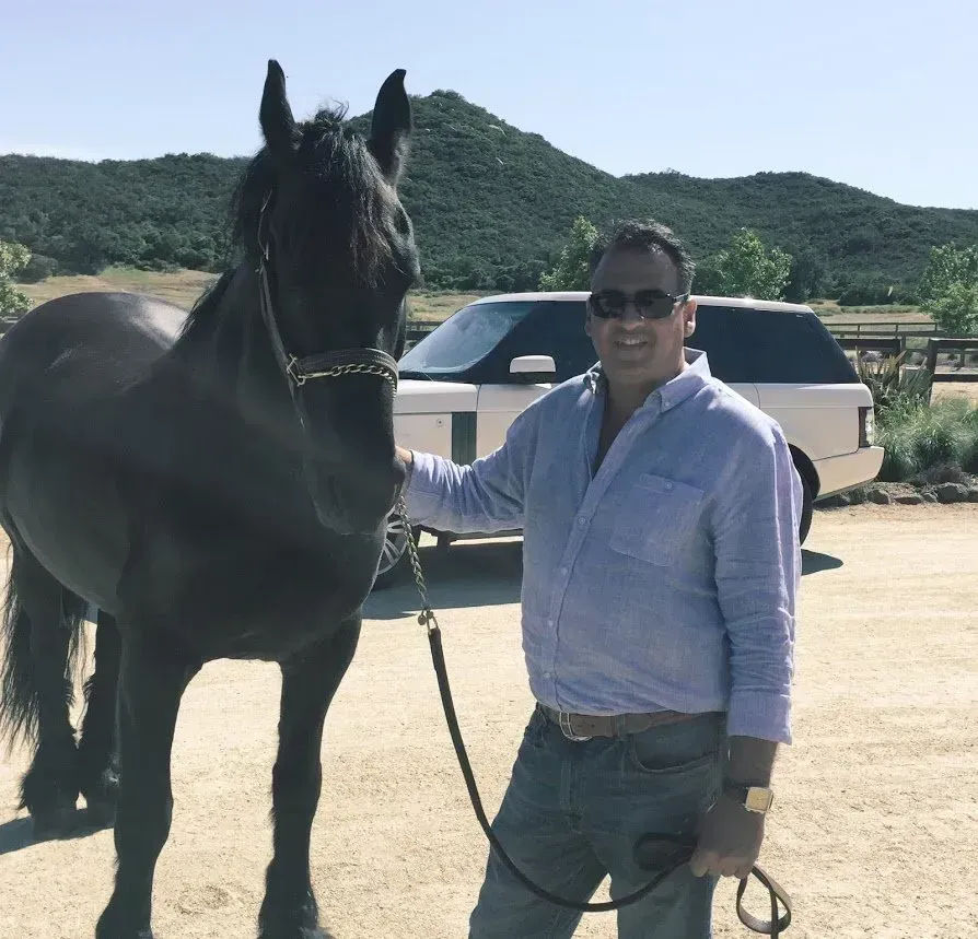 Man in sunglasses with black horse, holding a lead rope, in front of a white SUV and hills.