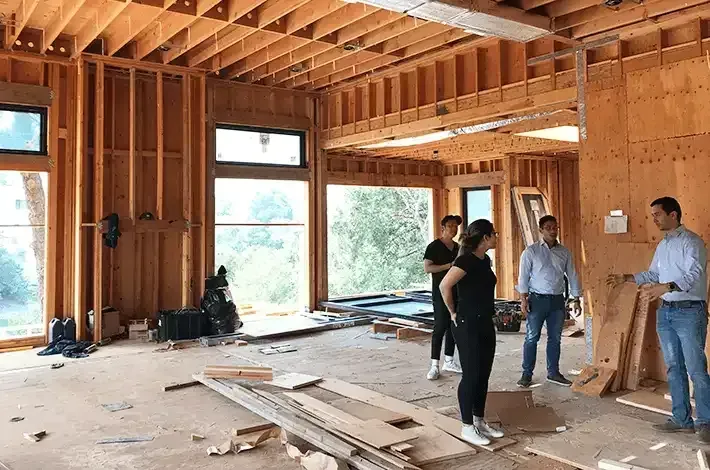 People in a wood-framed room during construction, discussing plans. Windows offer a view.
