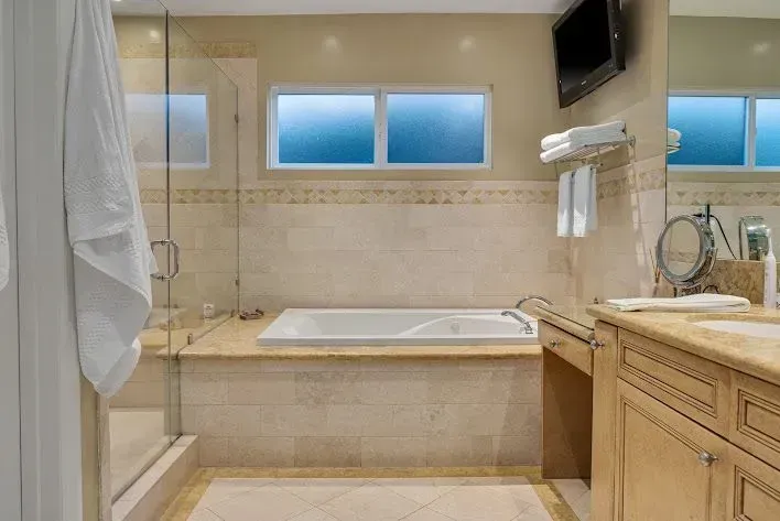 Bathroom with beige tile, a tub, shower, sink, window, and small TV. Towels are hanging.