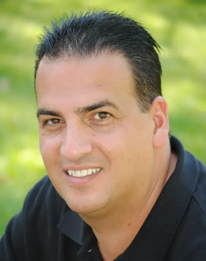 Man with dark hair and a black shirt smiling outdoors.