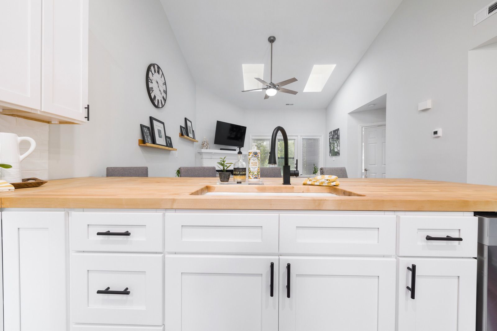 White kitchen with wooden countertop, black faucet, and hardware, looking into a living space.