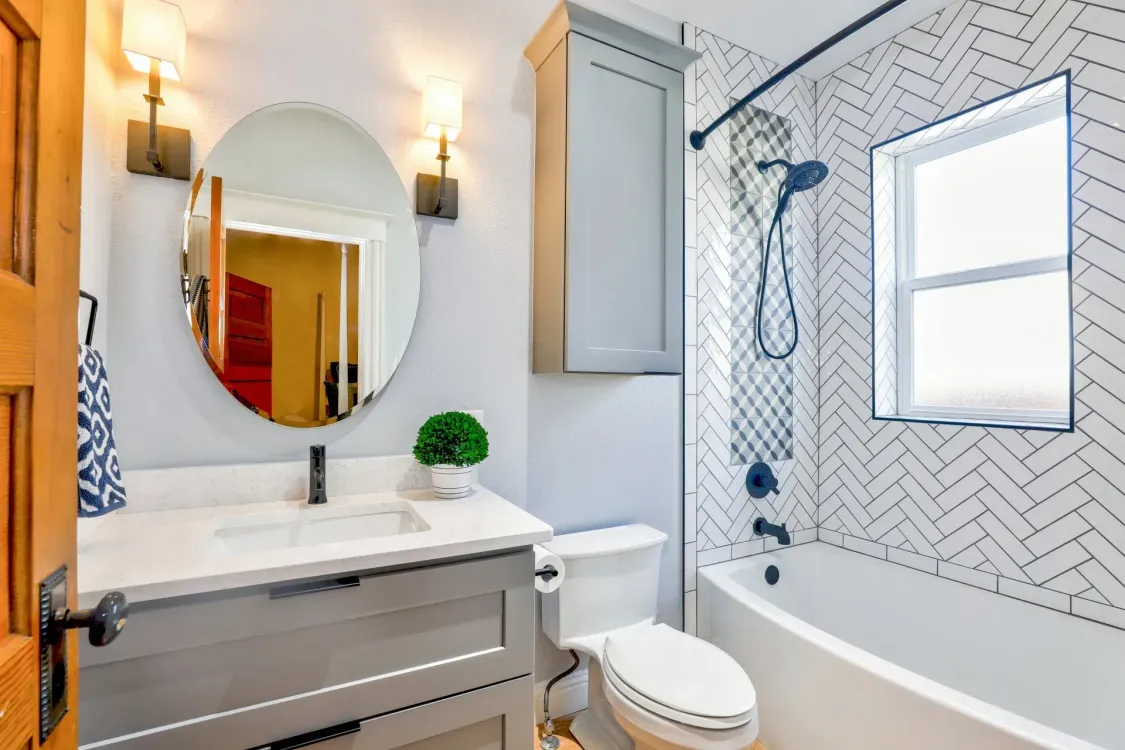 Bathroom with white and gray walls, round mirror, vanity, toilet, and bathtub with patterned tile.