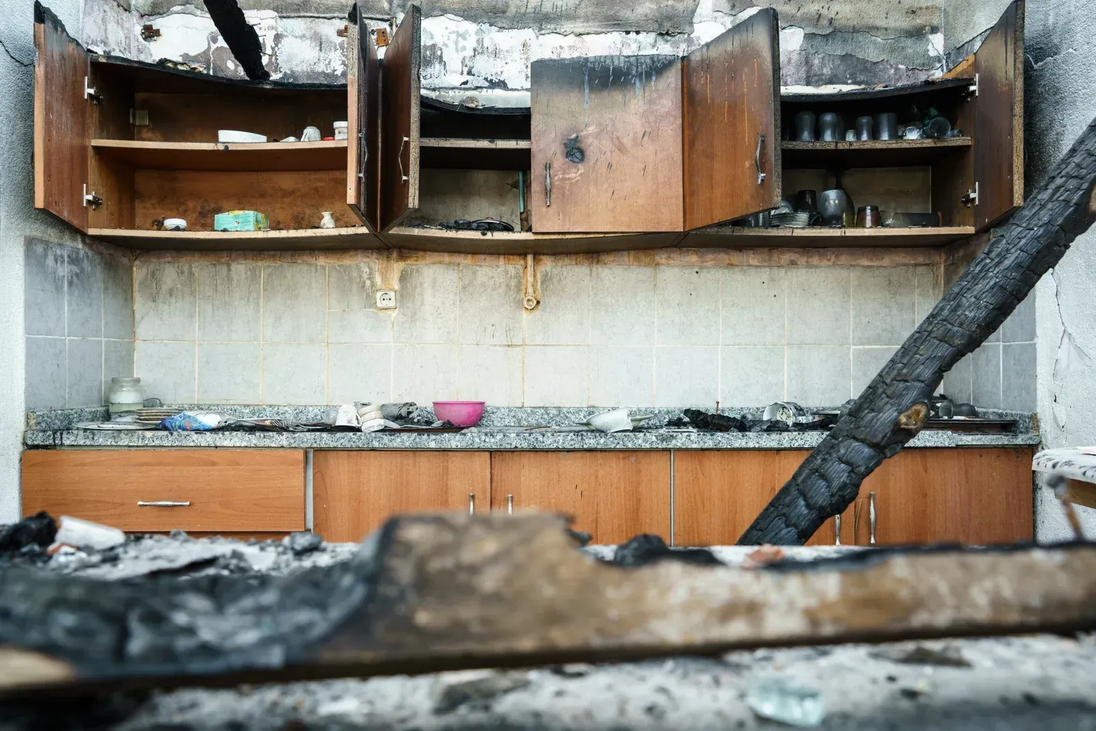 Burnt kitchen interior. Soot covers walls, cabinets open, charred beam and debris in foreground.