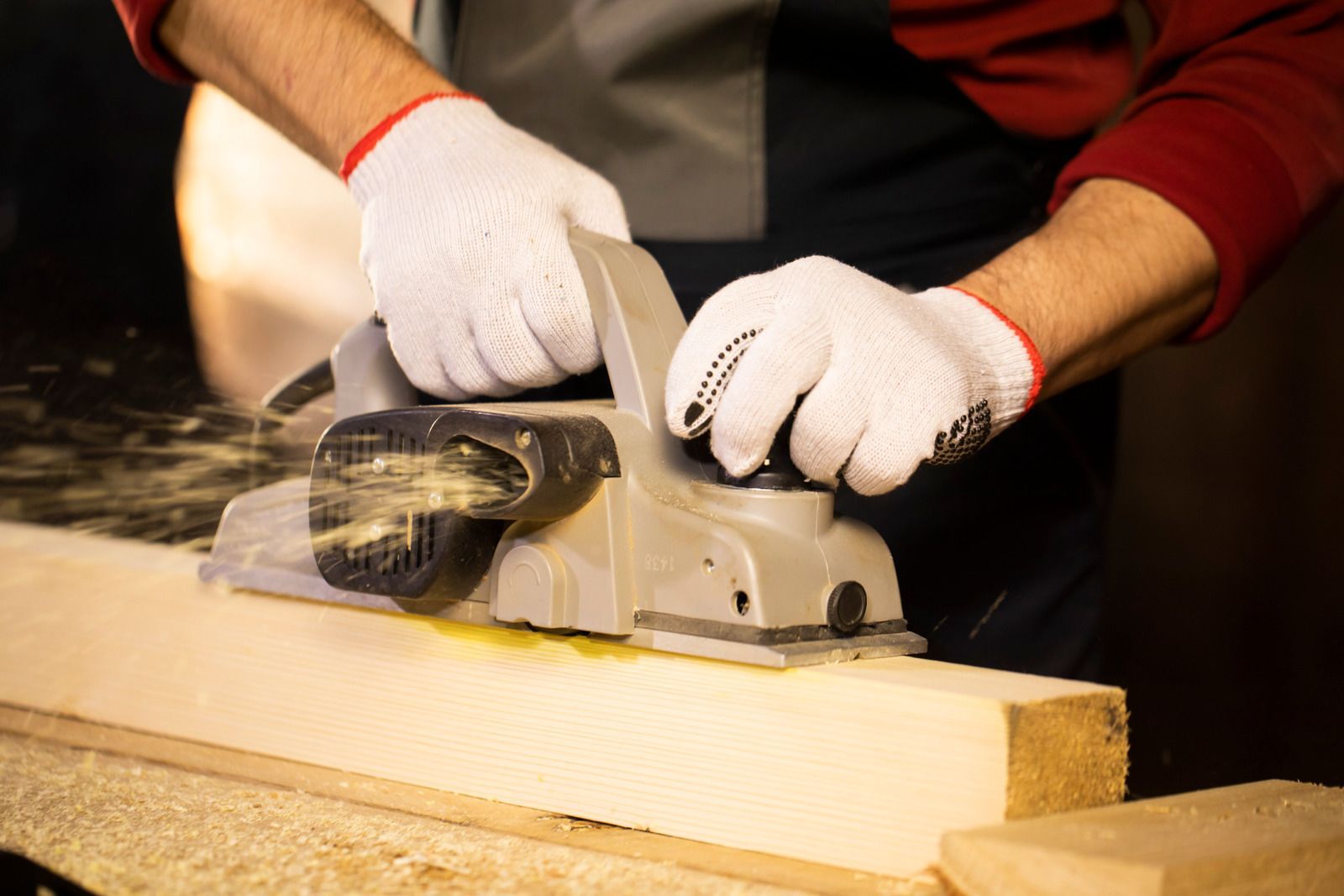 Person using a power planer to smooth a wooden board, wearing white gloves.