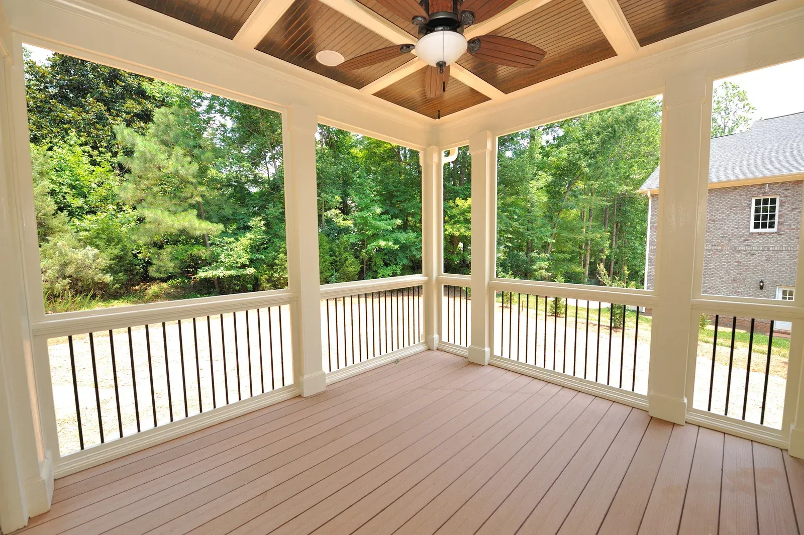 Screened porch with brown wooden floor, white columns, and black railings, overlooking green trees.