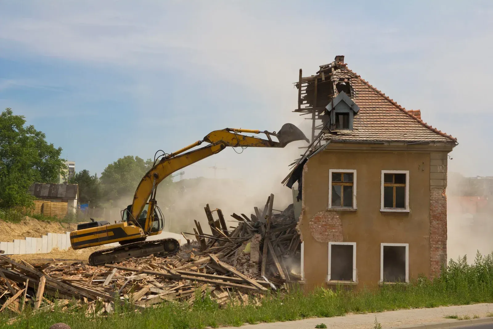 Excavator demolishing a two-story house, creating dust; sunny day.