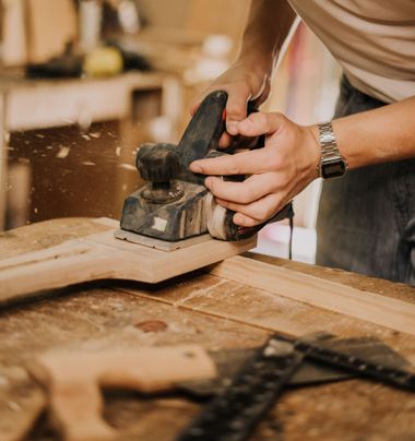 Person sanding a wooden board with a power sander in a workshop, with wood dust flying.