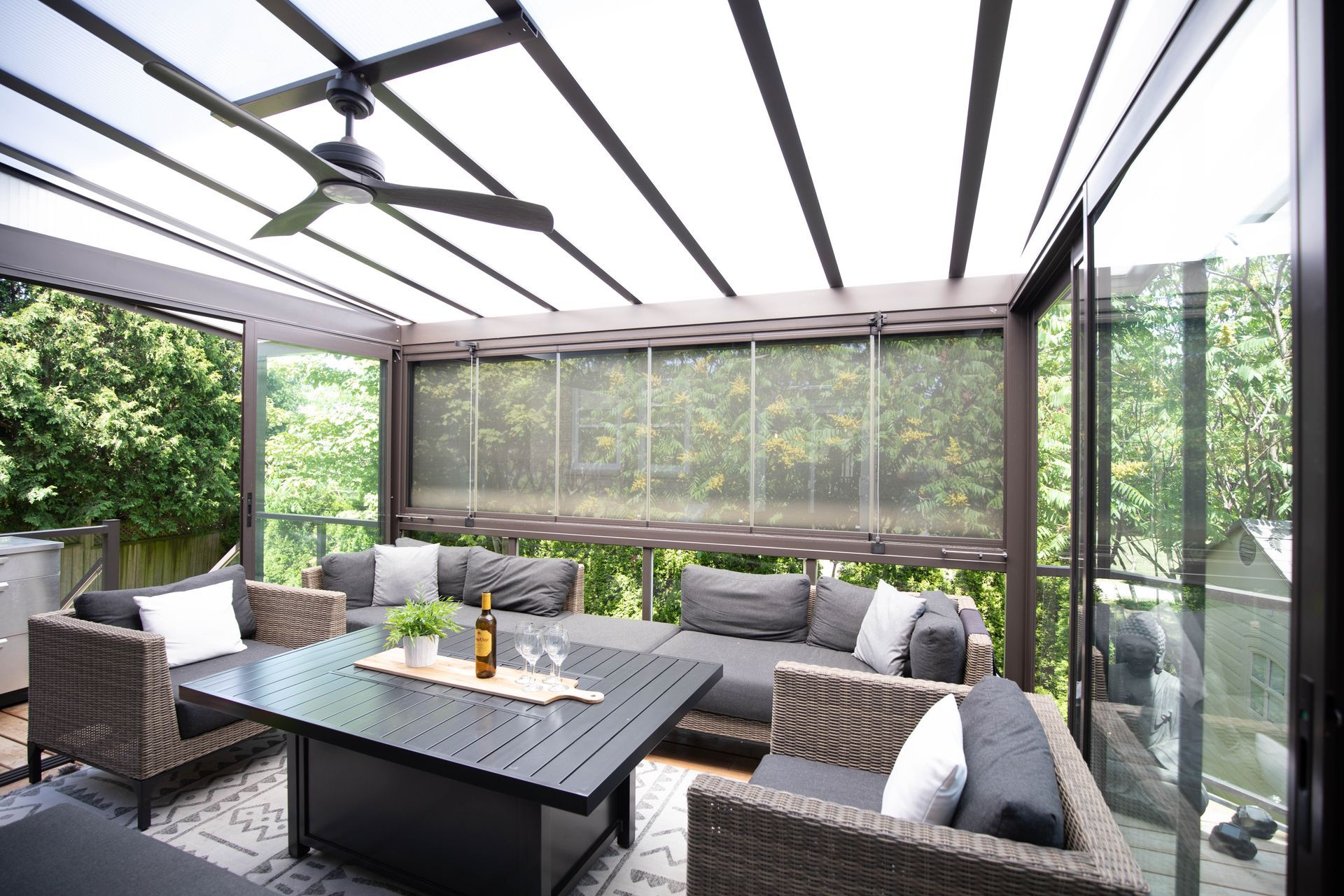 Outdoor seating area with modern furniture, glass walls, and a slatted roof, surrounded by trees.
