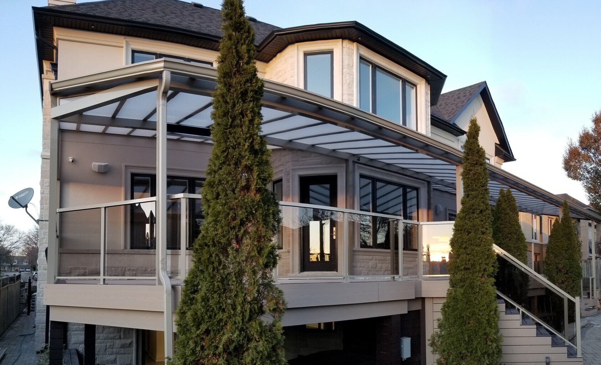 Two-story house with a deck and transparent roof. Three tall trees on the deck. Glass railings and black door.