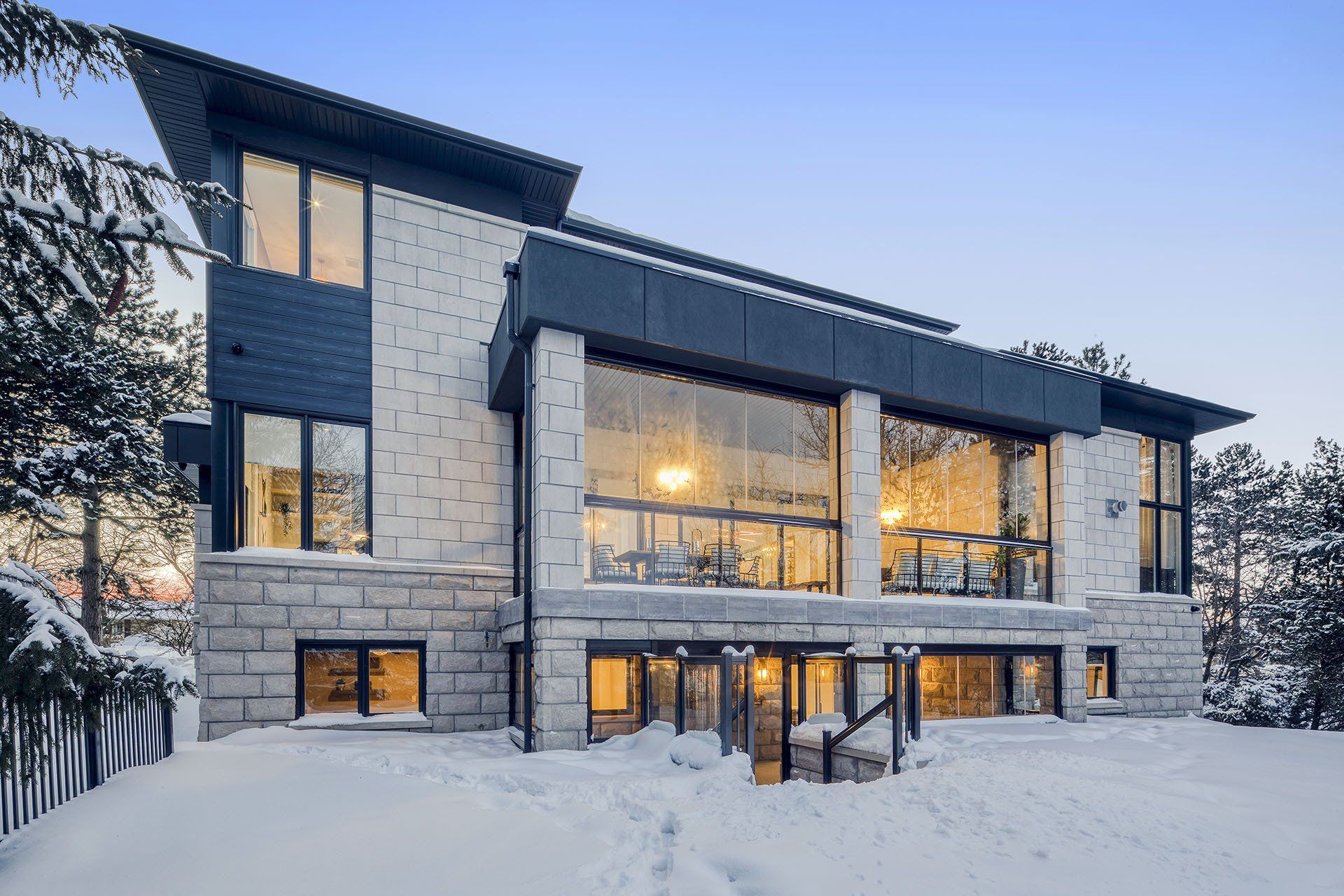 Modern house with large windows, stone facade, snow-covered ground, and dusk lighting.