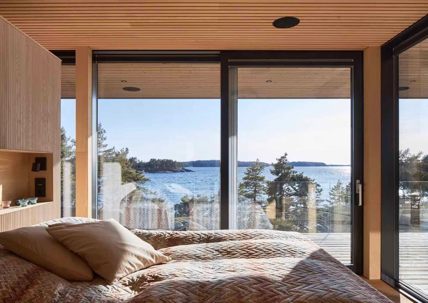 Bedroom with large windows overlooking a seascape. Wood-paneled walls, bed with pillows.