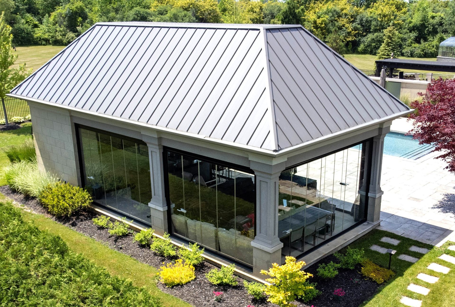Gazebo with glass walls and a metal roof near a pool. Surrounded by green grass and landscaping.