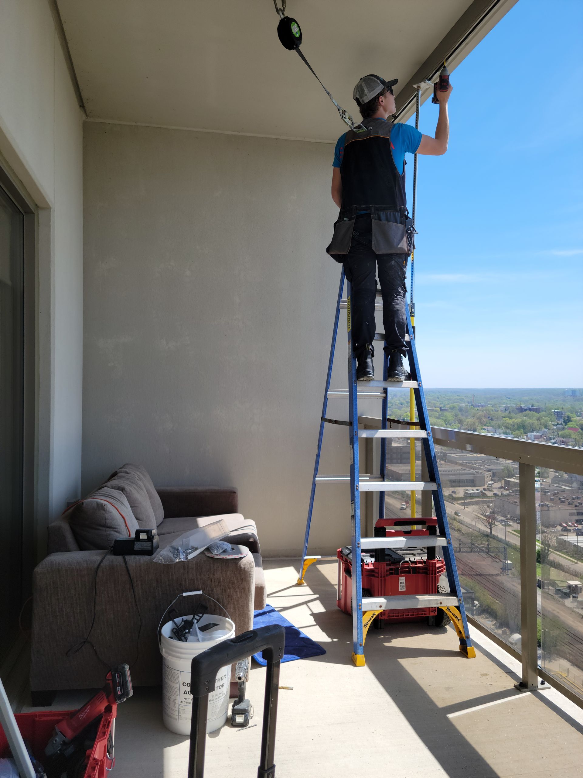 Person on a ladder cleaning a high-rise balcony window. Blue sky, cityscape, and furniture in the background.