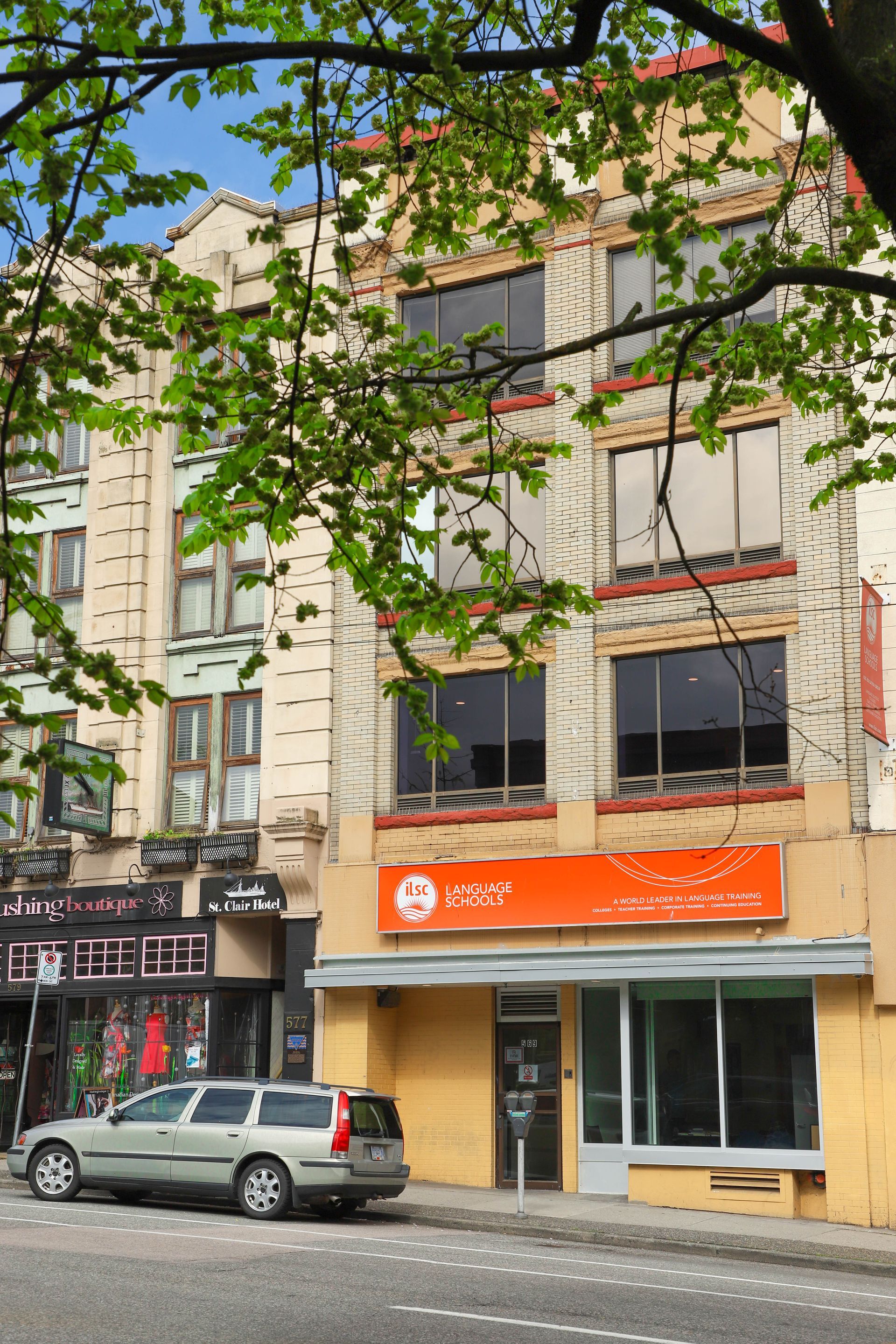 Street-level view of a brick storefront with orange awning, parked car, and leafy branches in front