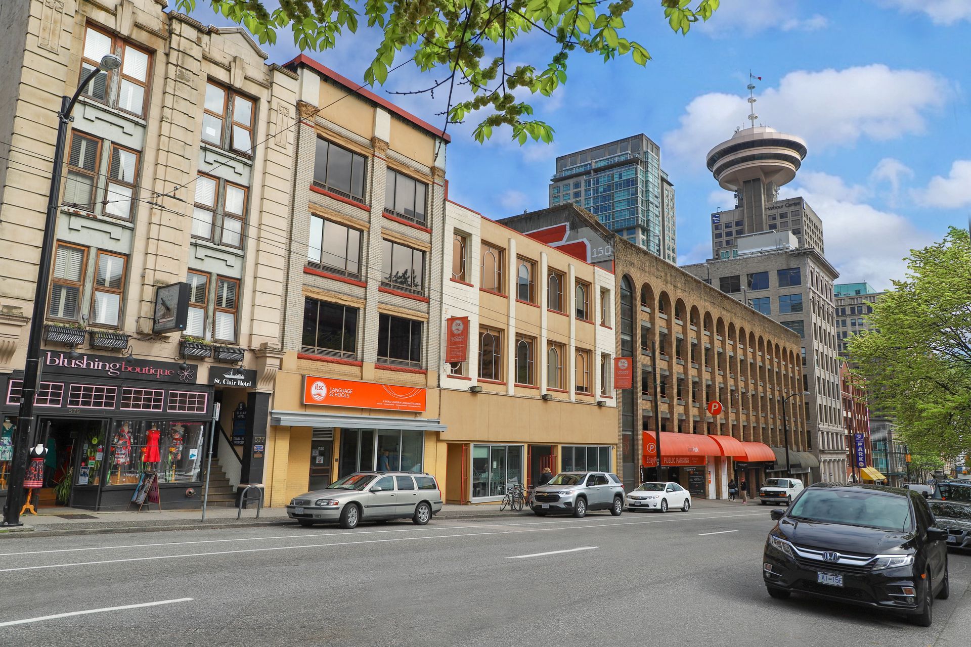 Urban street with shops, parked cars, trees, and a distinctive tower in the background under a blue sky