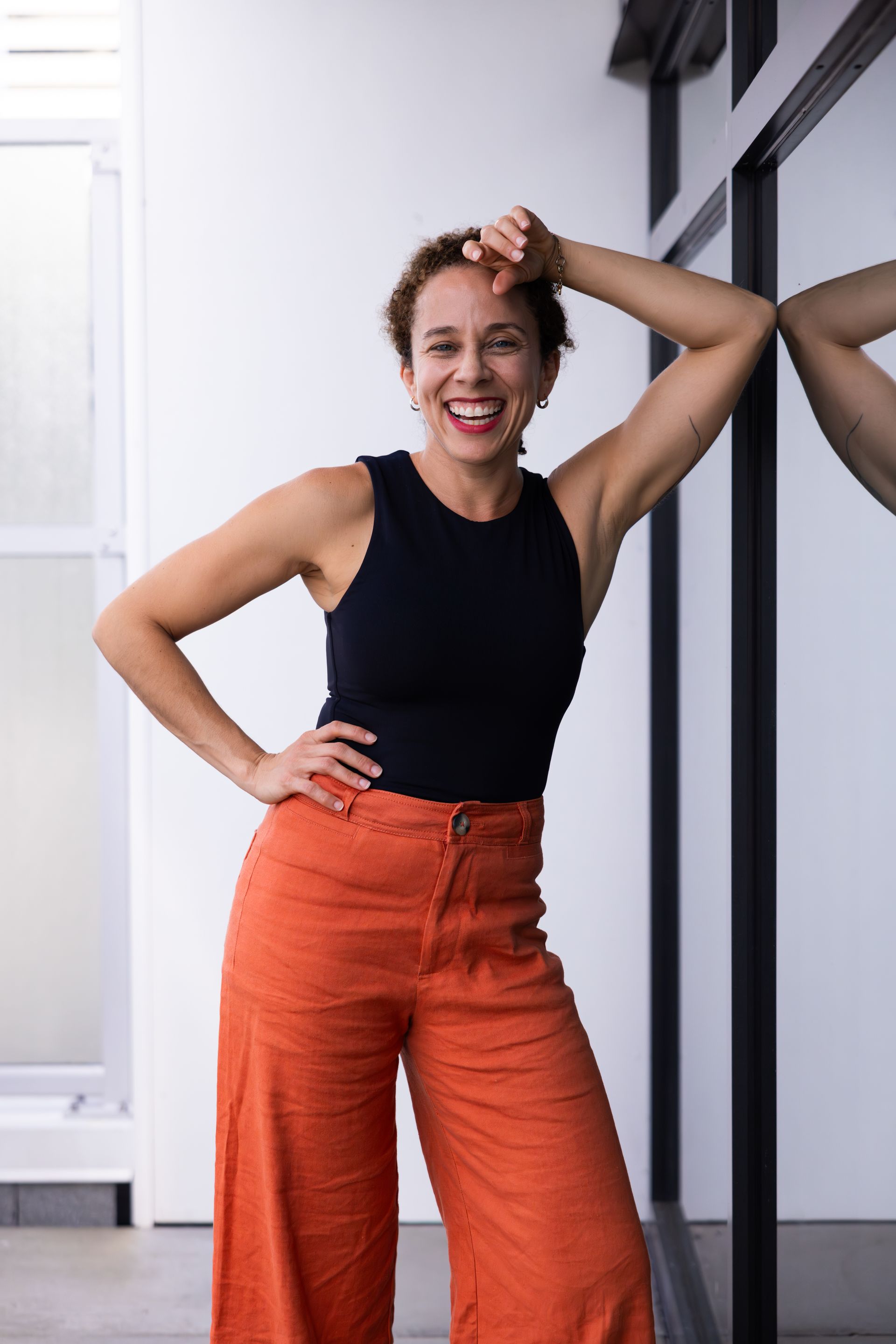 Woman in black top and orange pants smiling and posing by a wall mirror