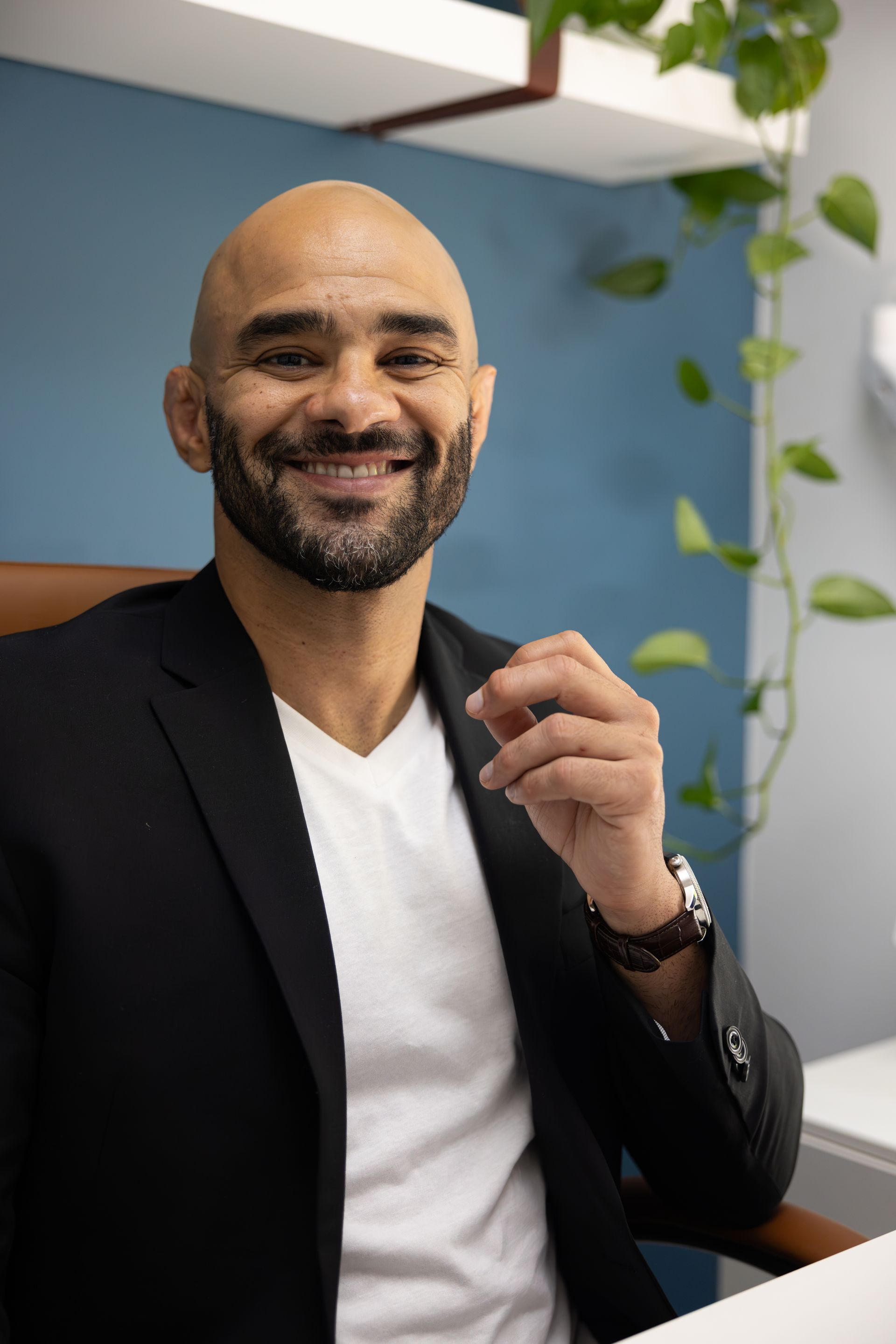 Smiling man in a black blazer and white shirt seated indoors against a blue wall with hanging plants