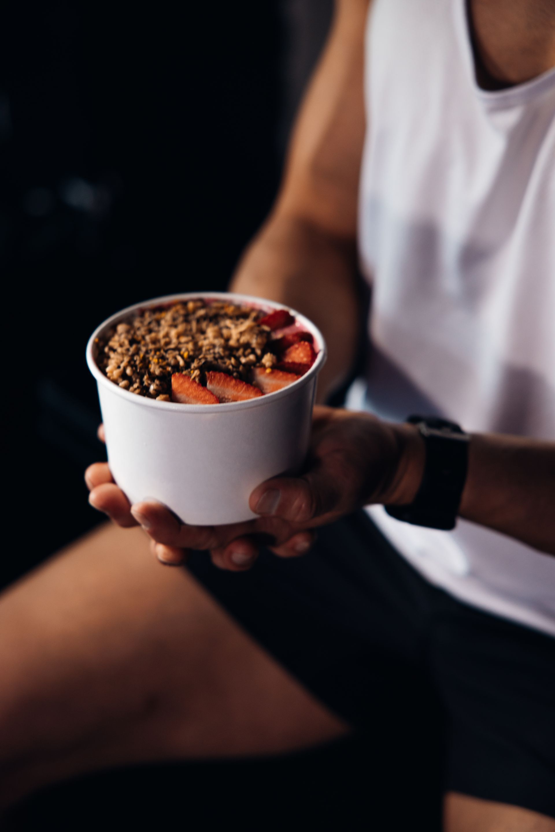Person holding a white bowl of granola and strawberries in a dim indoor setting