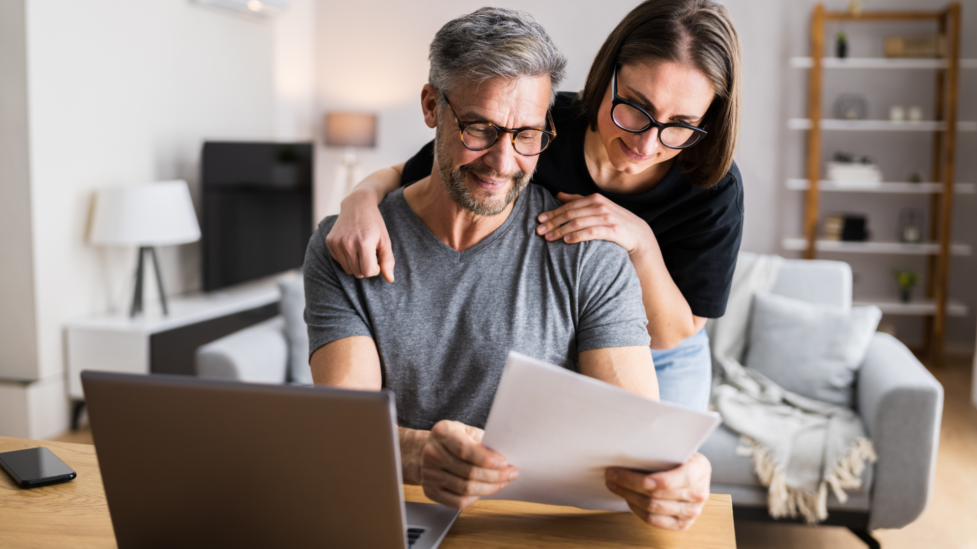 Two people review papers together beside a laptop in a bright living room.