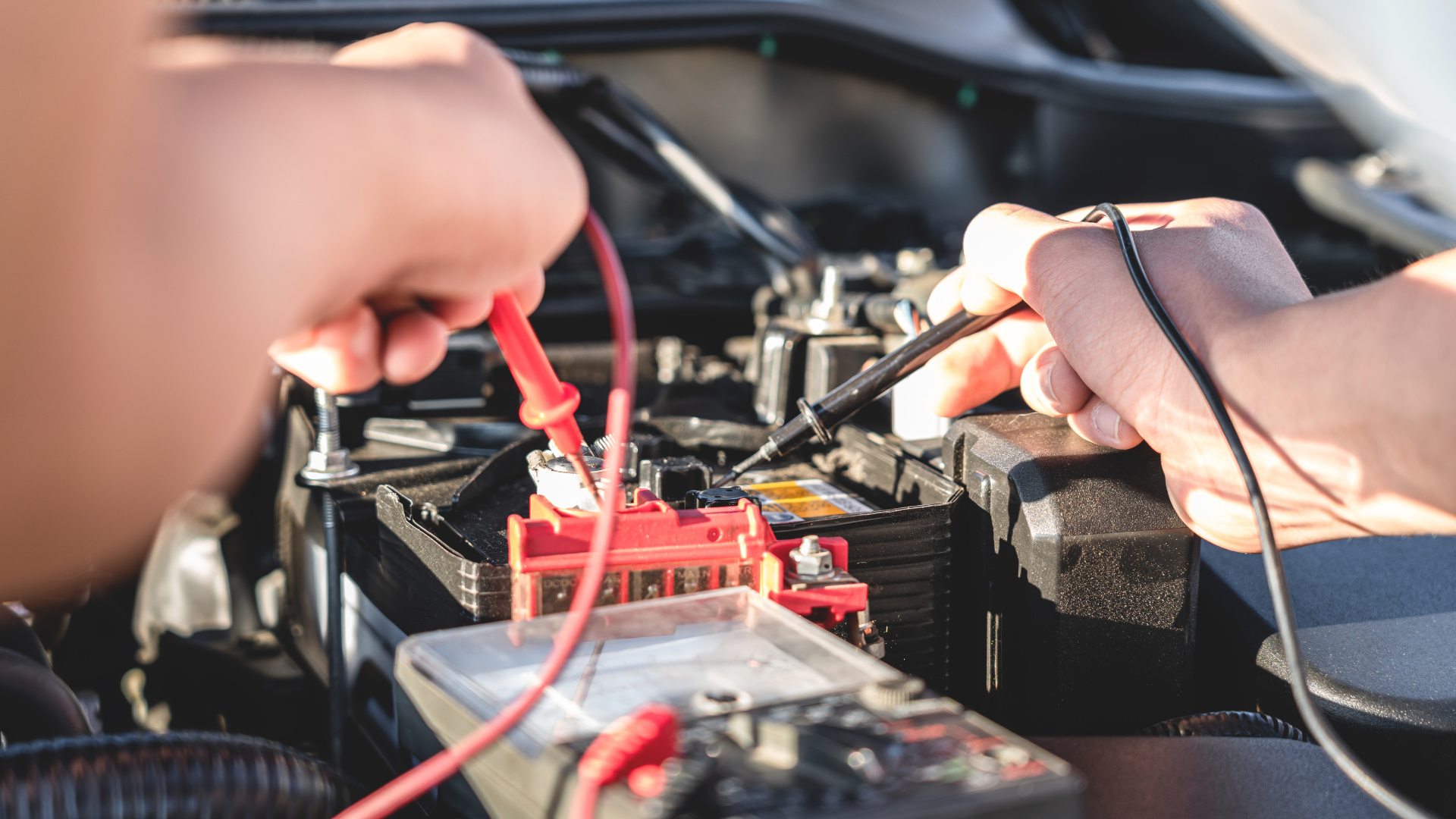 Hands use a multimeter with red and black probes to test a car battery inside an engine bay.