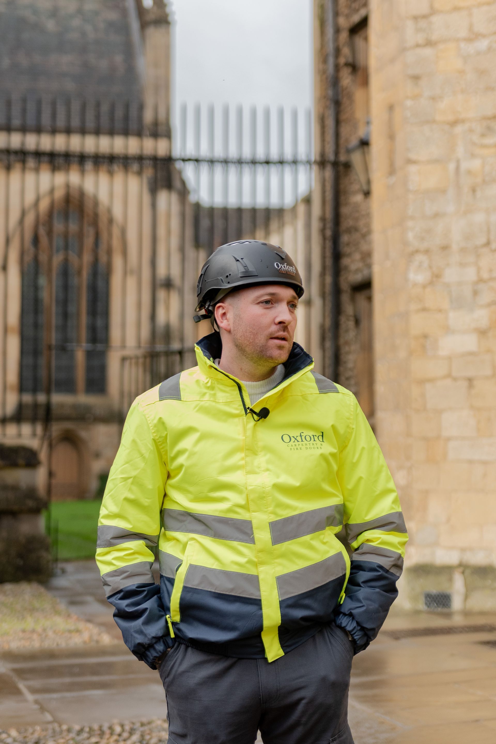 Man in safety vest and helmet standing near a stone building and gate, looking off to the side.