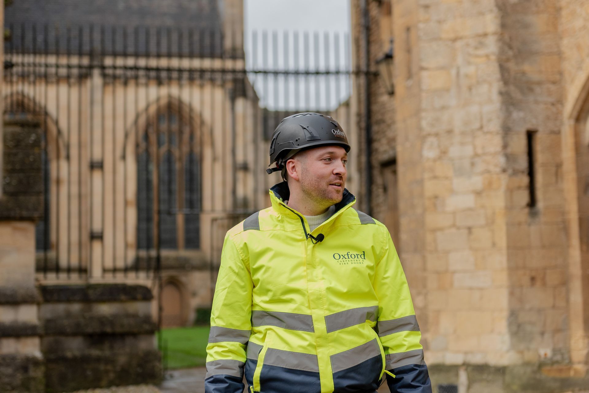 Man in safety gear, helmet, and high-vis jacket, near an arched stone building with a metal gate.