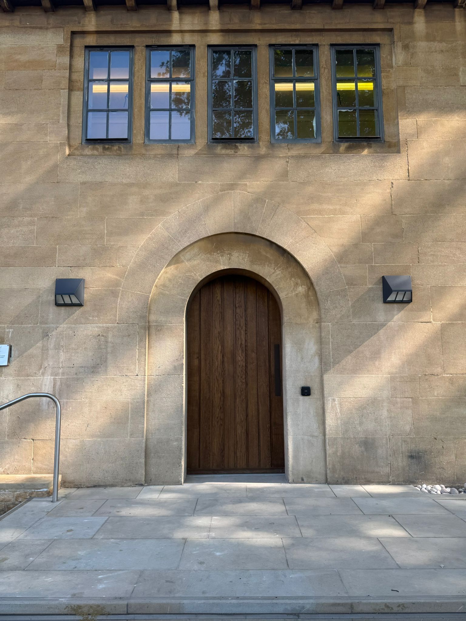 Stone building facade with arched wooden door, three windows above, two wall sconces.