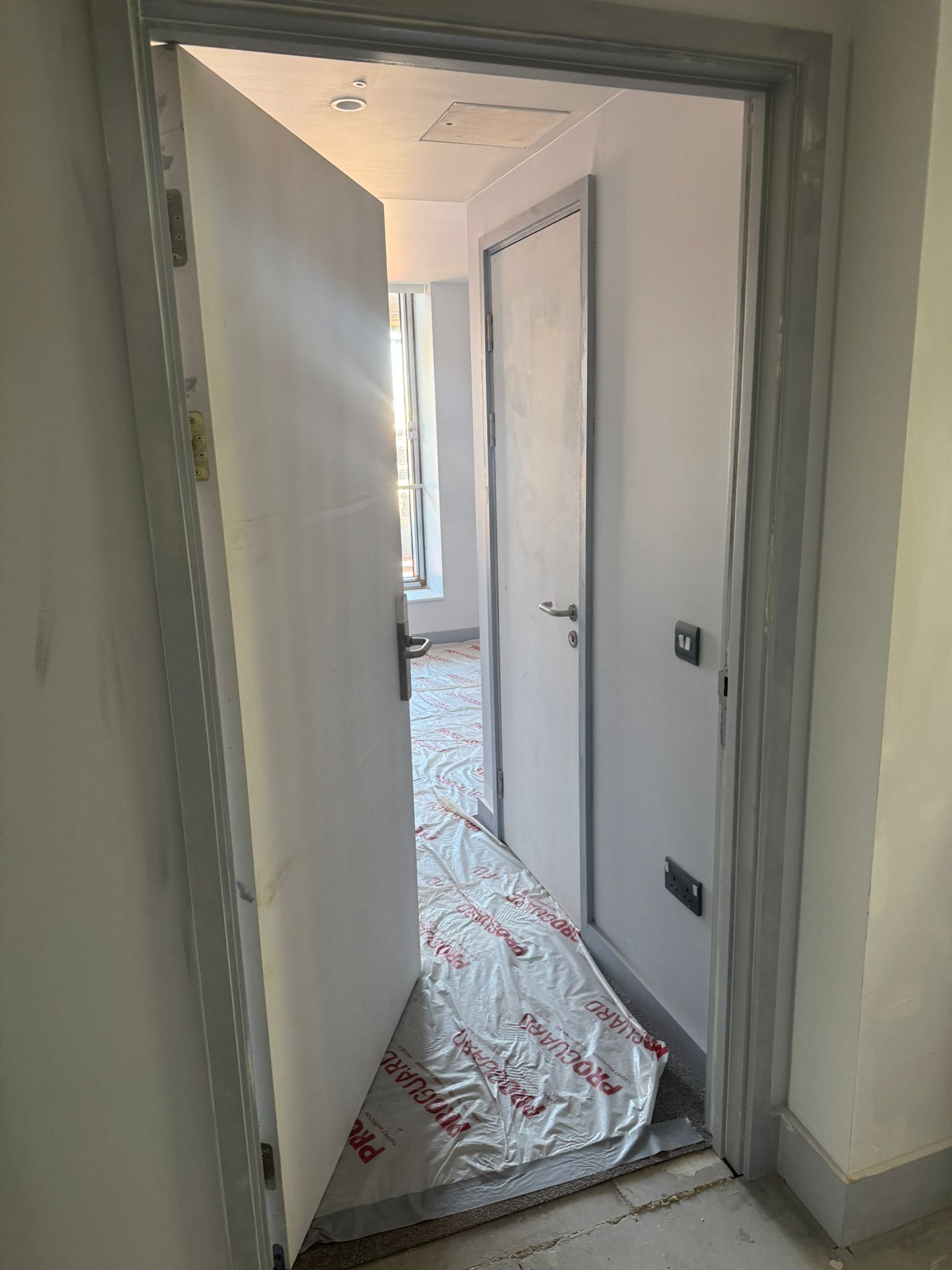 Open white door revealing a new hallway with another closed door, and protective floor covering.