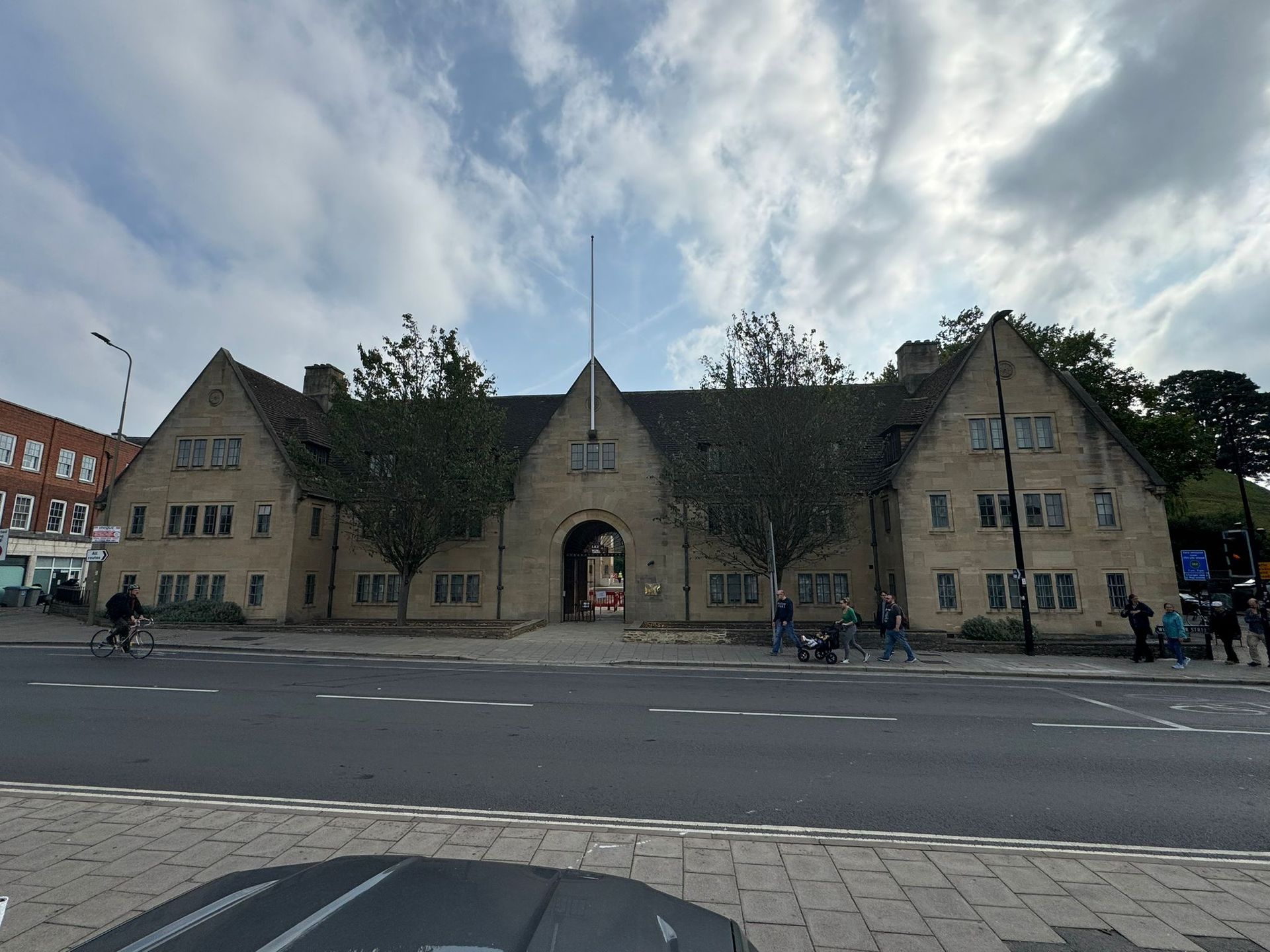 Stone building with archway entrance, multiple gabled roofs, trees, road, and cloudy sky.