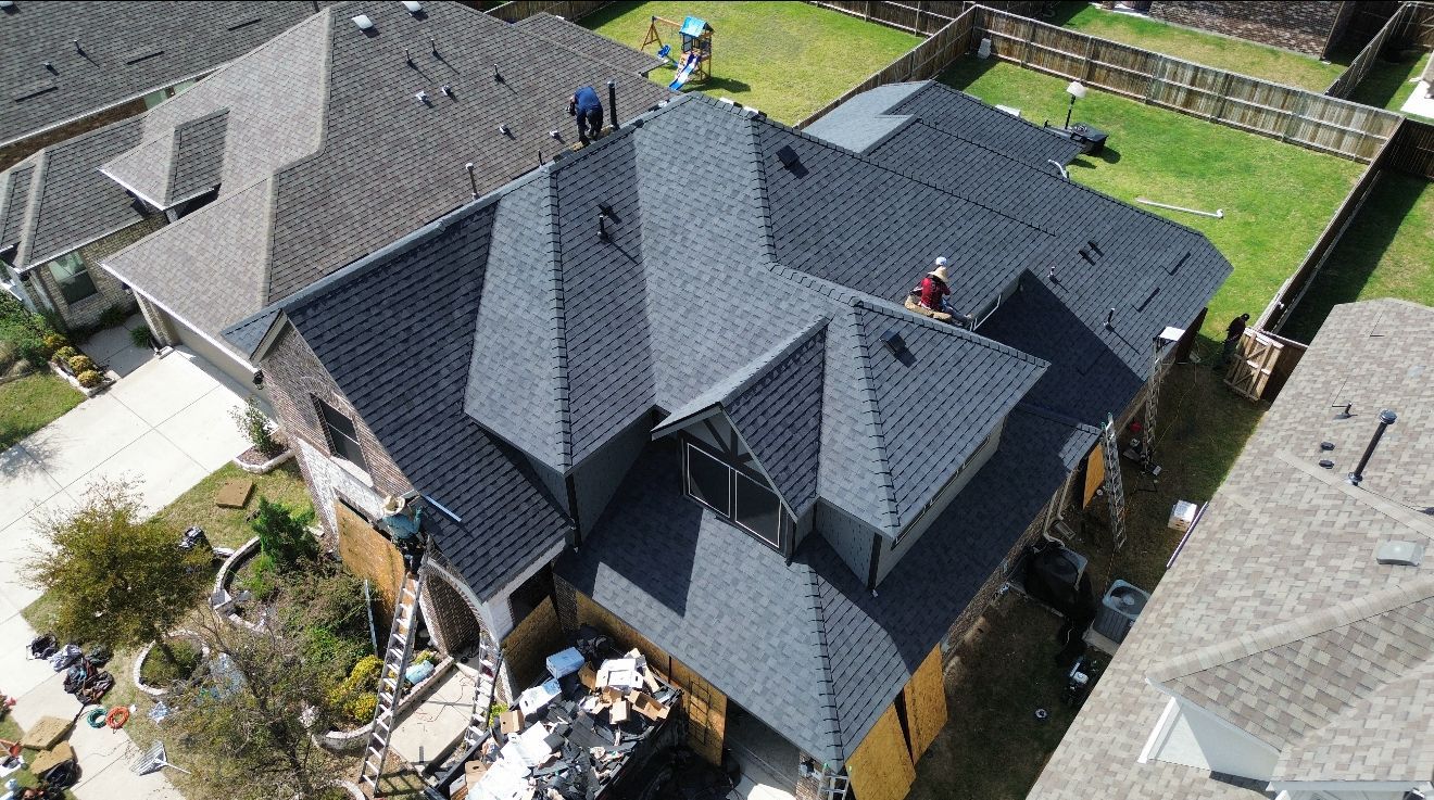 Aerial view of a house with a dark gray roof being worked on by two people. Green lawn in the backyard.
