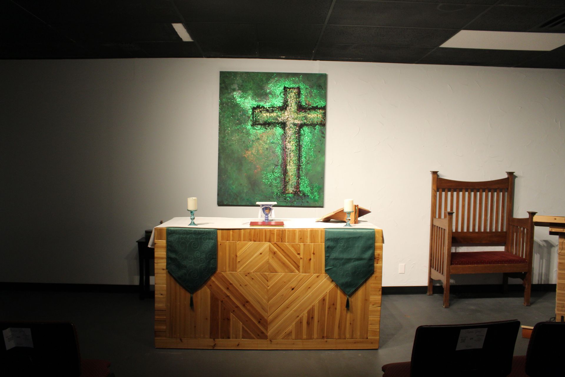Altar with a wooden cross, candles, and a chair. Green cross on green background. Light illuminates the altar.