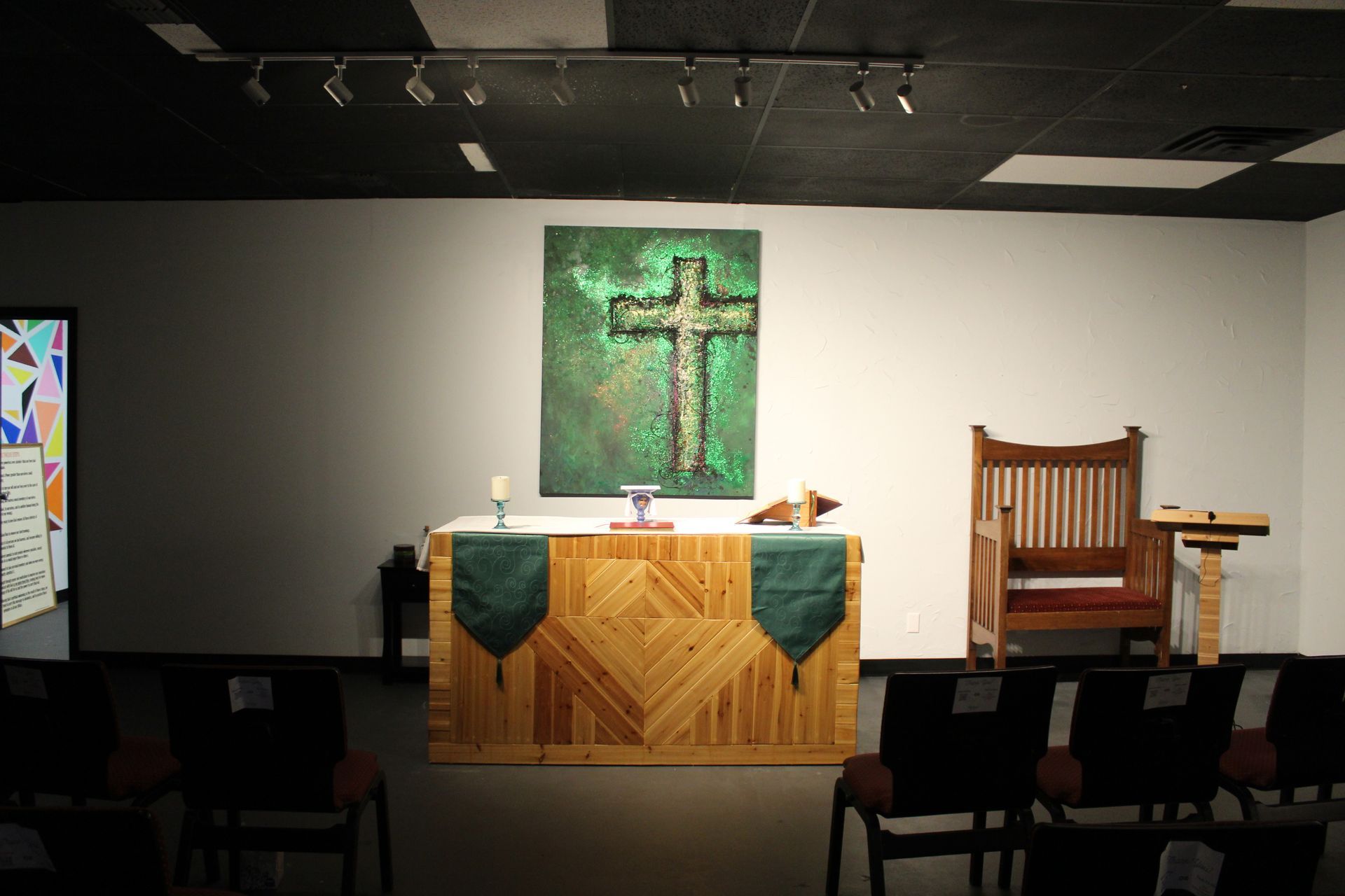 Altar with cross art, wooden furniture, and rows of chairs in a church. Green and brown tones.